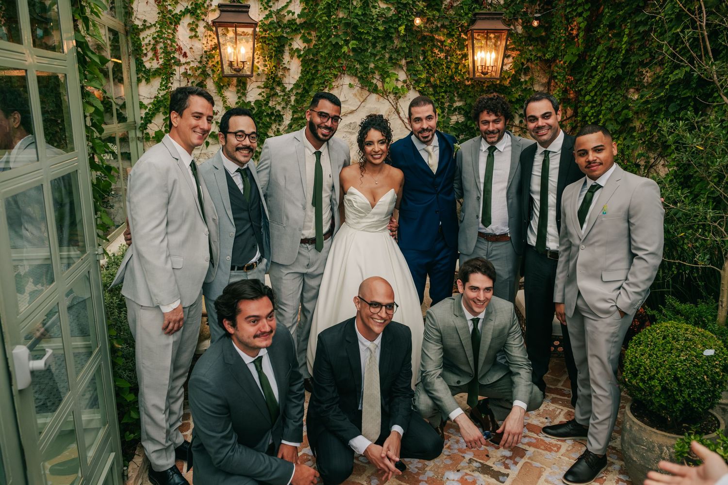 Bride and groom pose with groomsmen in grey suits at ivy-covered outdoor wedding venue.