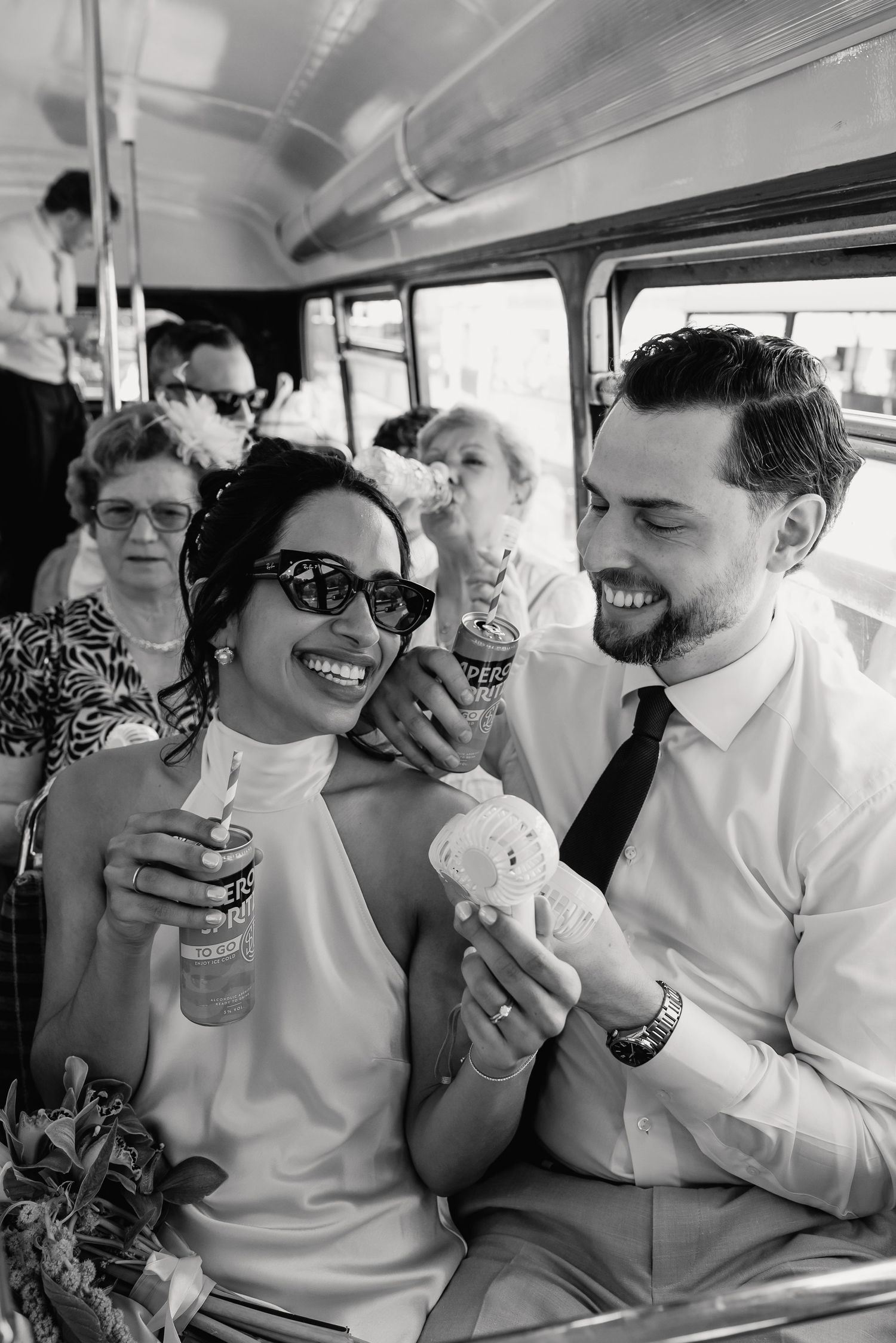 Bride and groom laughing on a vintage bus, holding ice cream and drinks, with wedding guests in background, black and white photo.