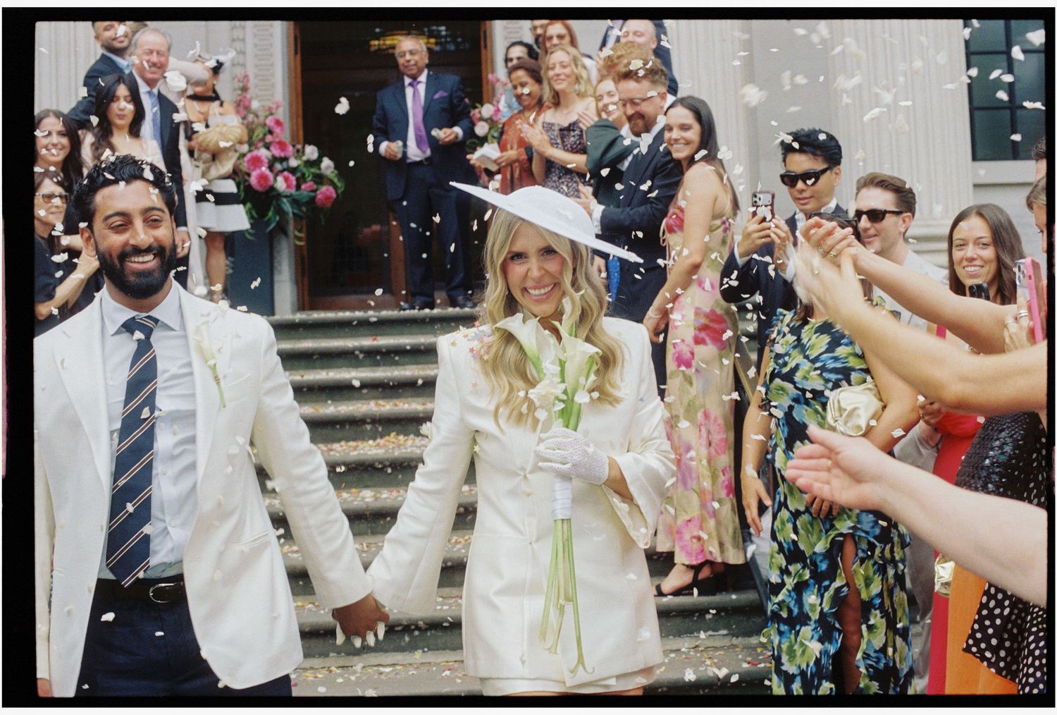 Newlyweds walk through confetti shower outside elegant building surrounded by cheering wedding guests.