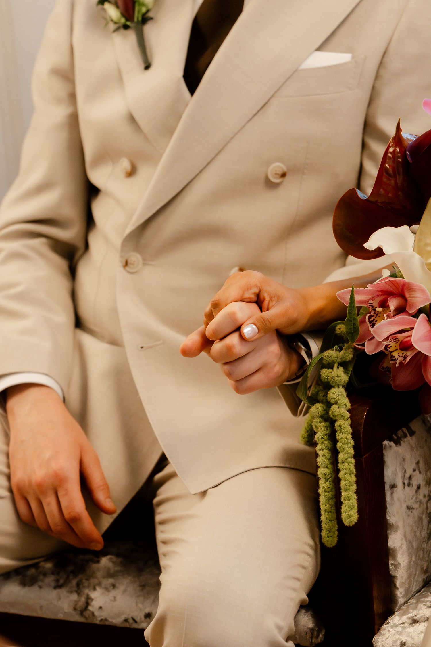 Groom in a beige double-breasted suit holds brides hand with a floral bouquet nearby during wedding ceremony.