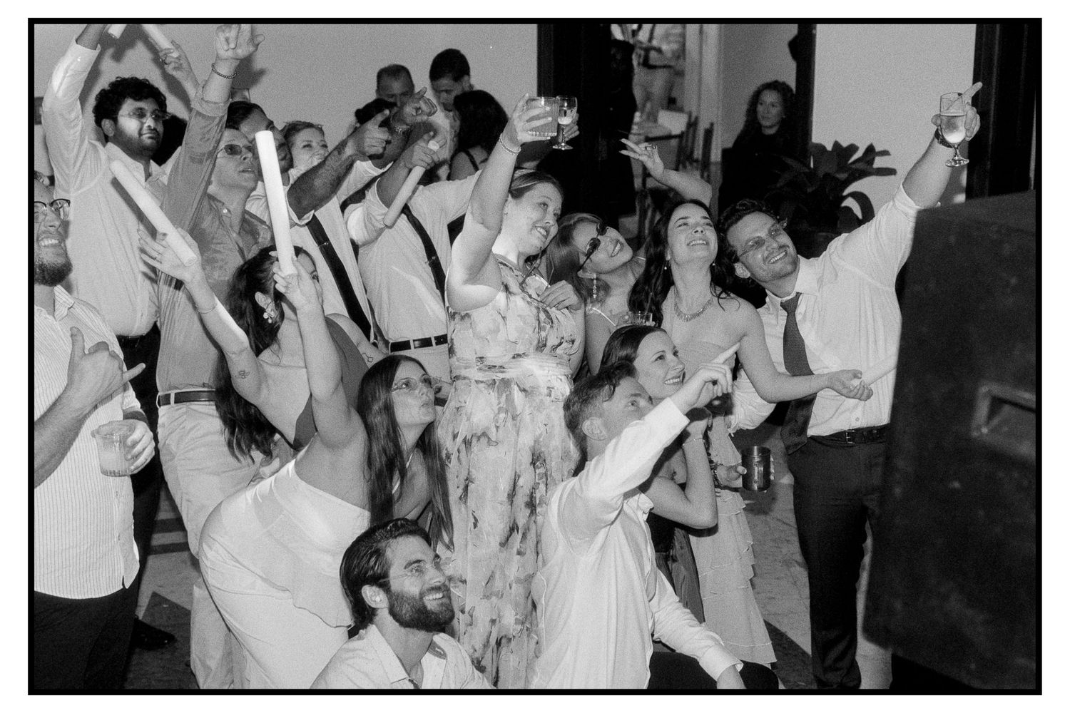 Black and white photo of large group of wedding guests celebrating with glow sticks on dancefloor.
