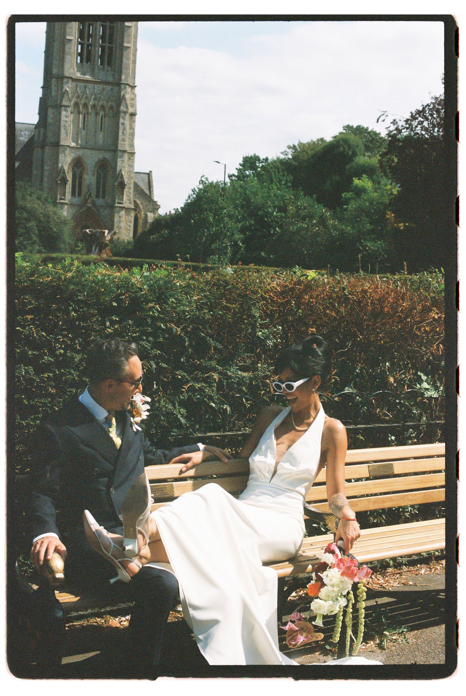 Newlyweds relax on a park bench with a historic church tower visible in the background.