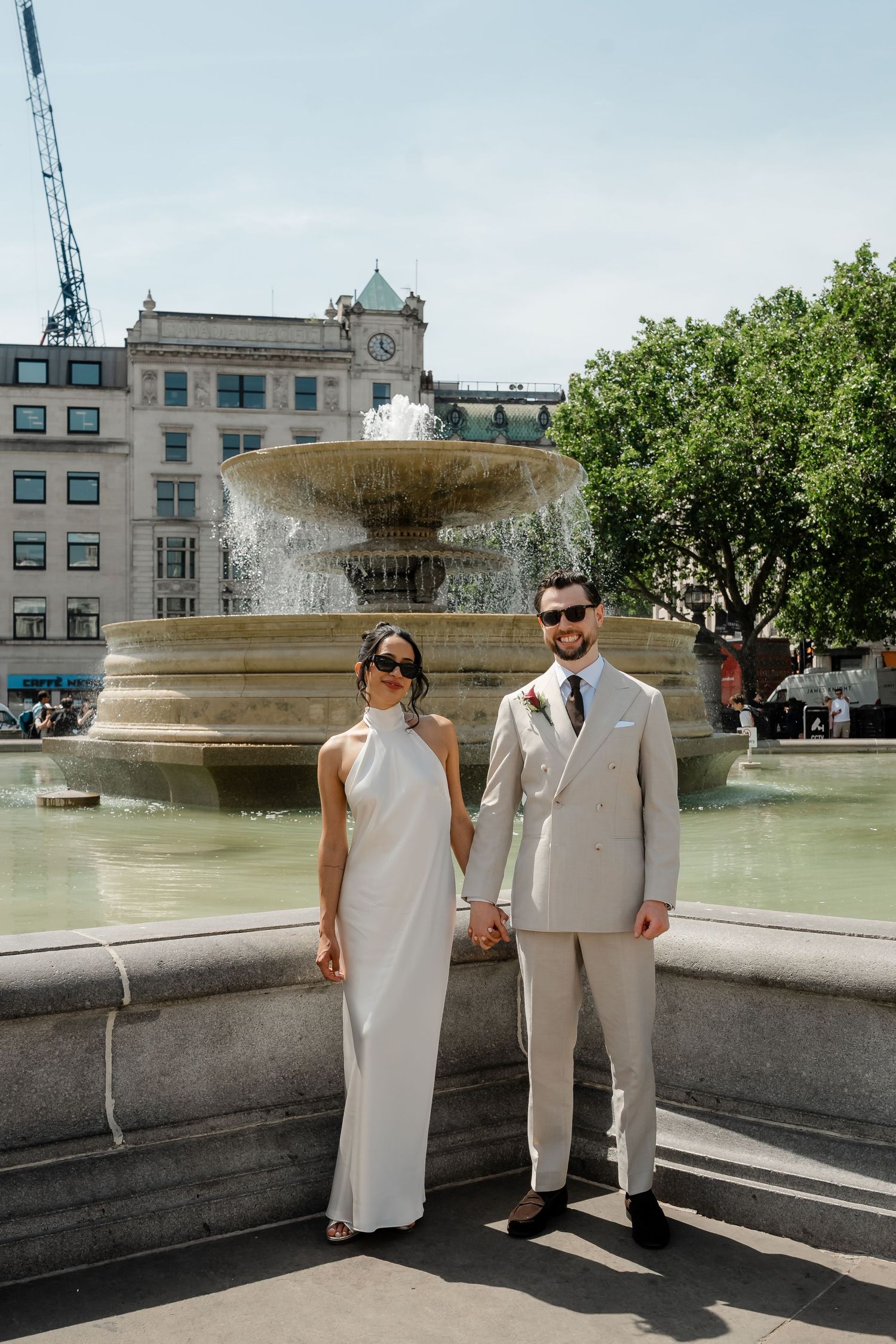 Elegant bride and groom in sunglasses hold hands before Trafalgar Square fountain in London.