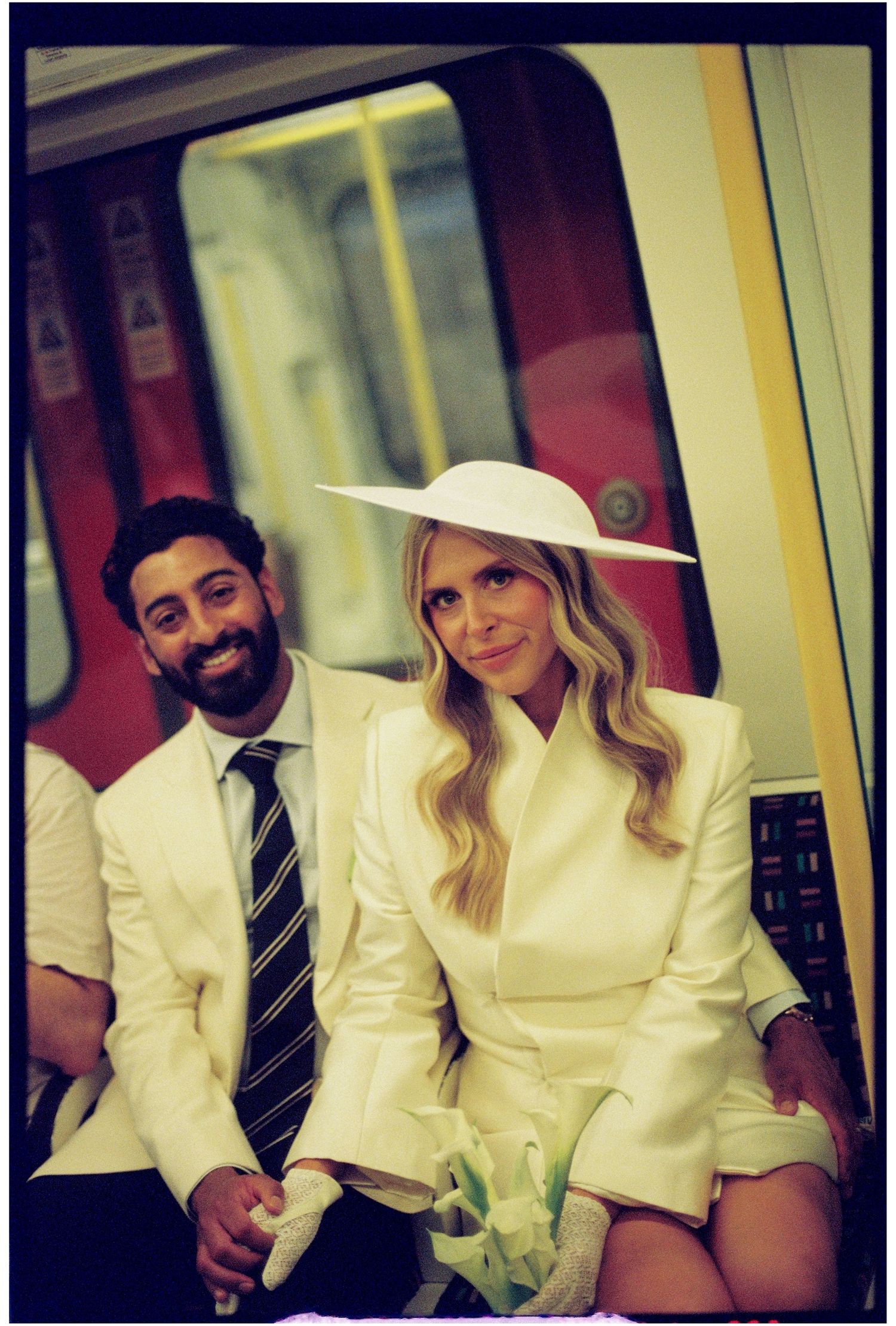 Stylish bride in white blazer and wide-brim hat sits with groom on London Underground train.