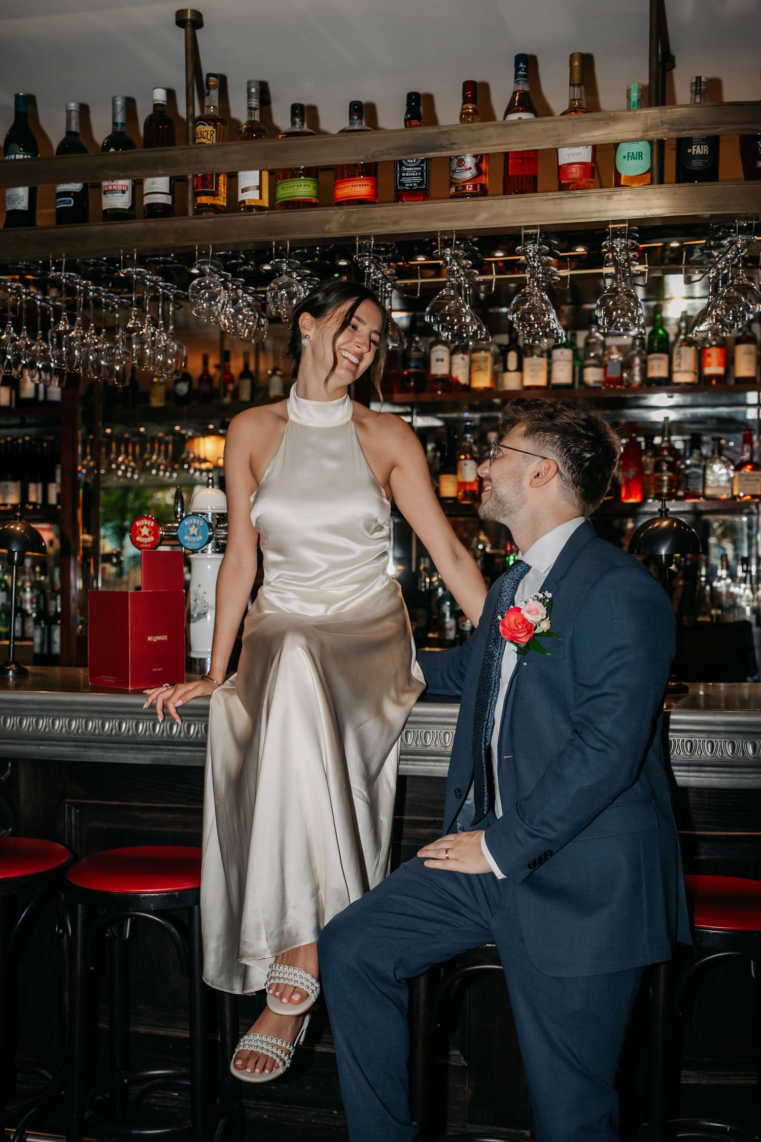 Bride in silver satin dress sits on bar counter laughing with groom in navy suit at elegant wedding venue.