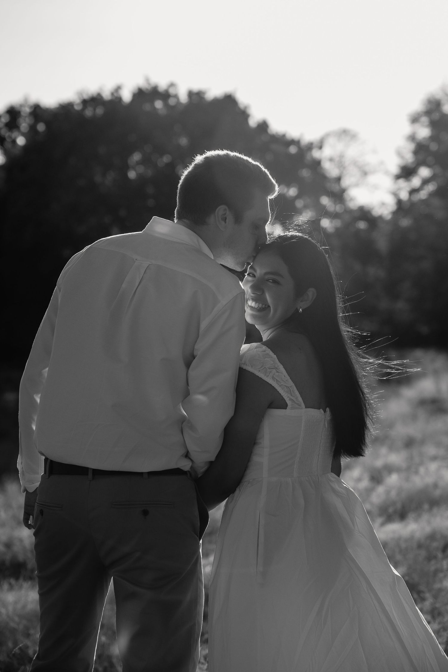 Black and white photo of a smiling bride in a white gown embracing a groom outdoors at sunset.