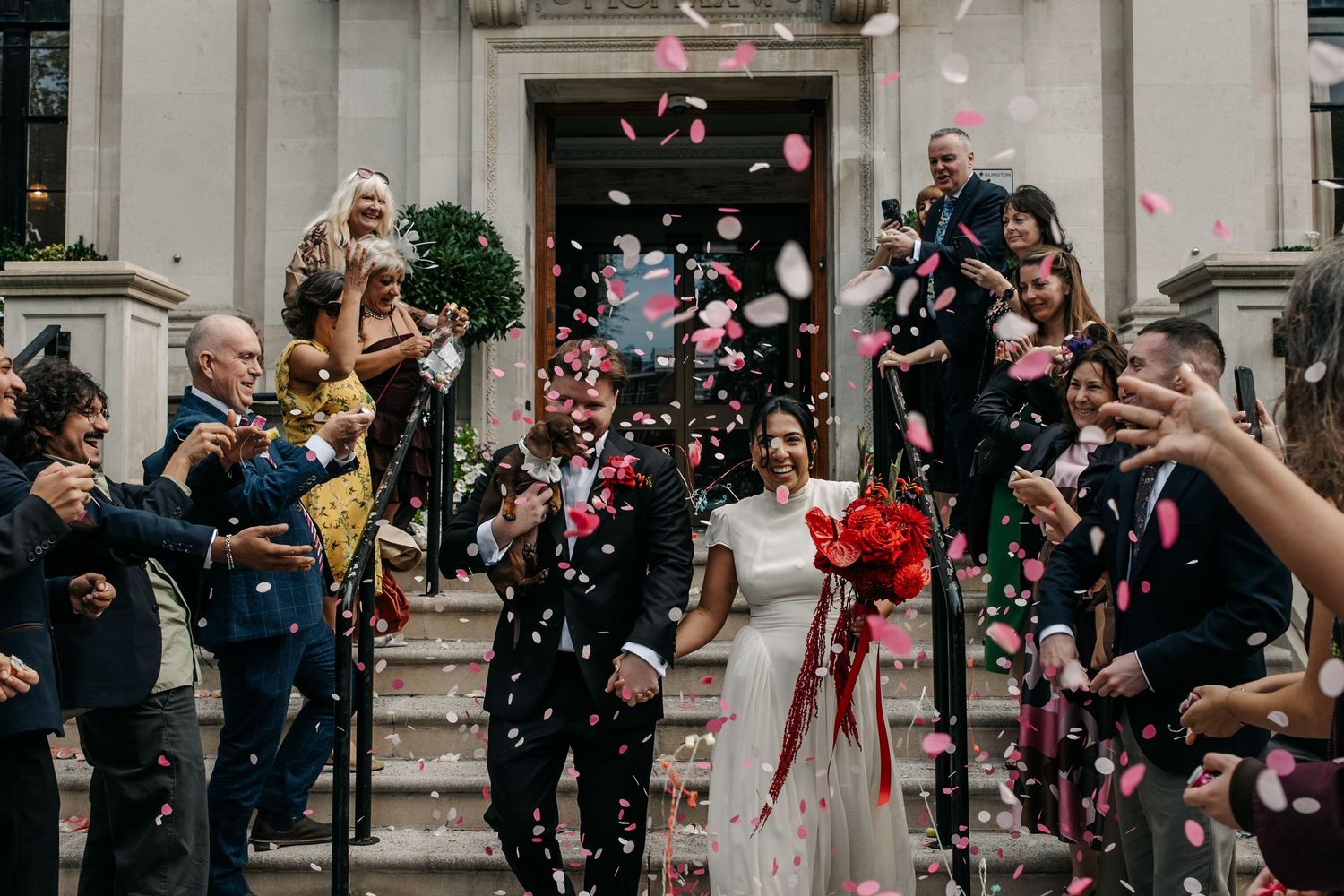 Joyful newlyweds walk through confetti shower outside elegant venue surrounded by cheering wedding guests.
