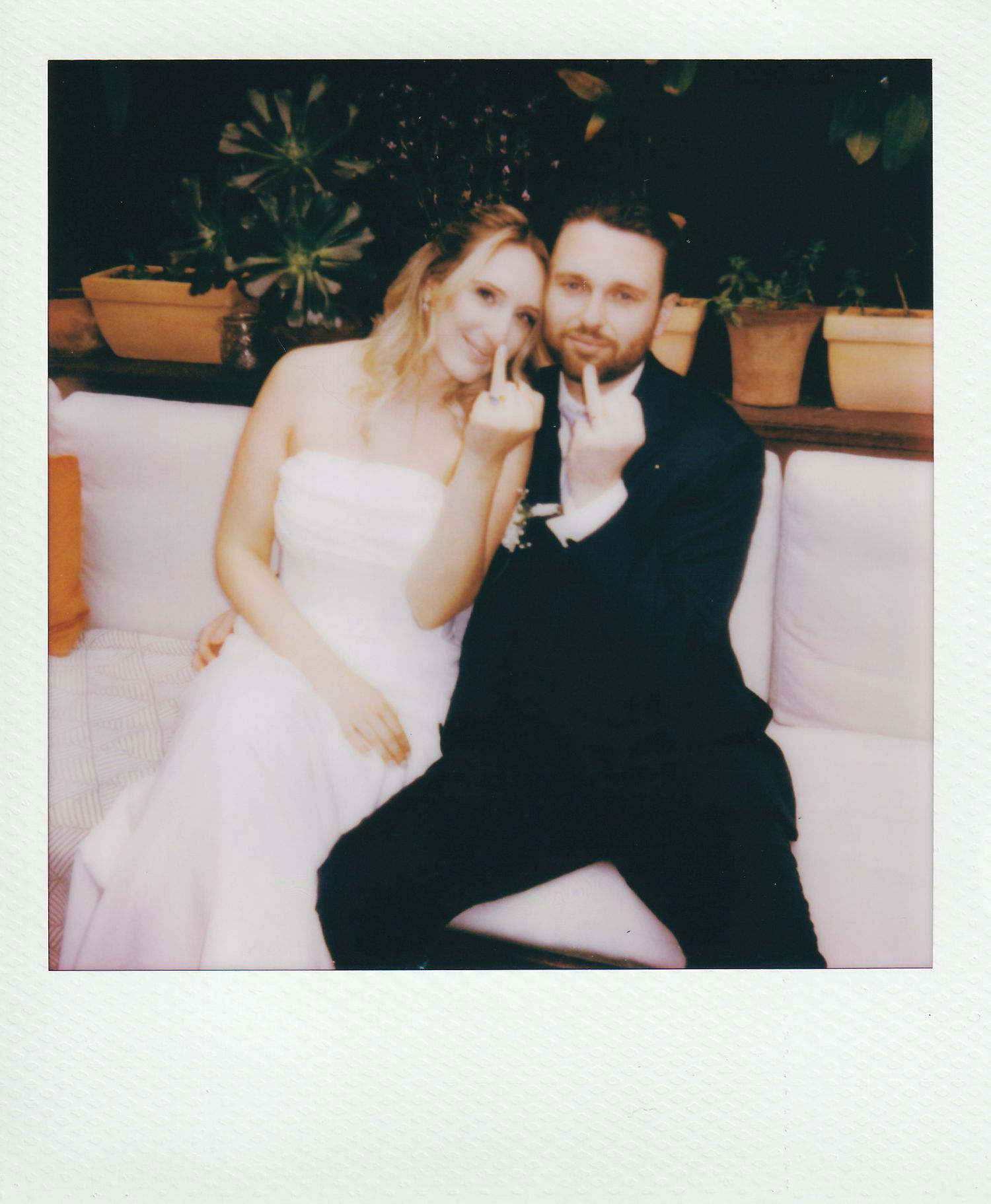 Polaroid photo of a bride and groom sitting together on a white couch, both making playful gestures at their wedding.