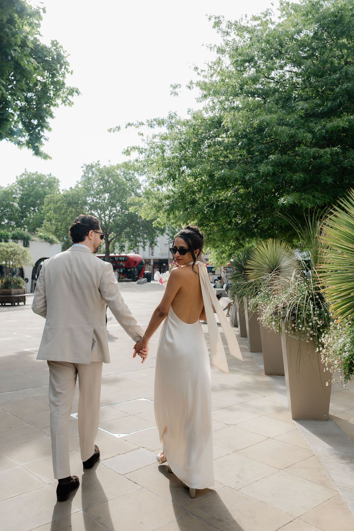 Bride in backless white gown and groom in beige suit holding hands walking along tree-lined urban plaza.