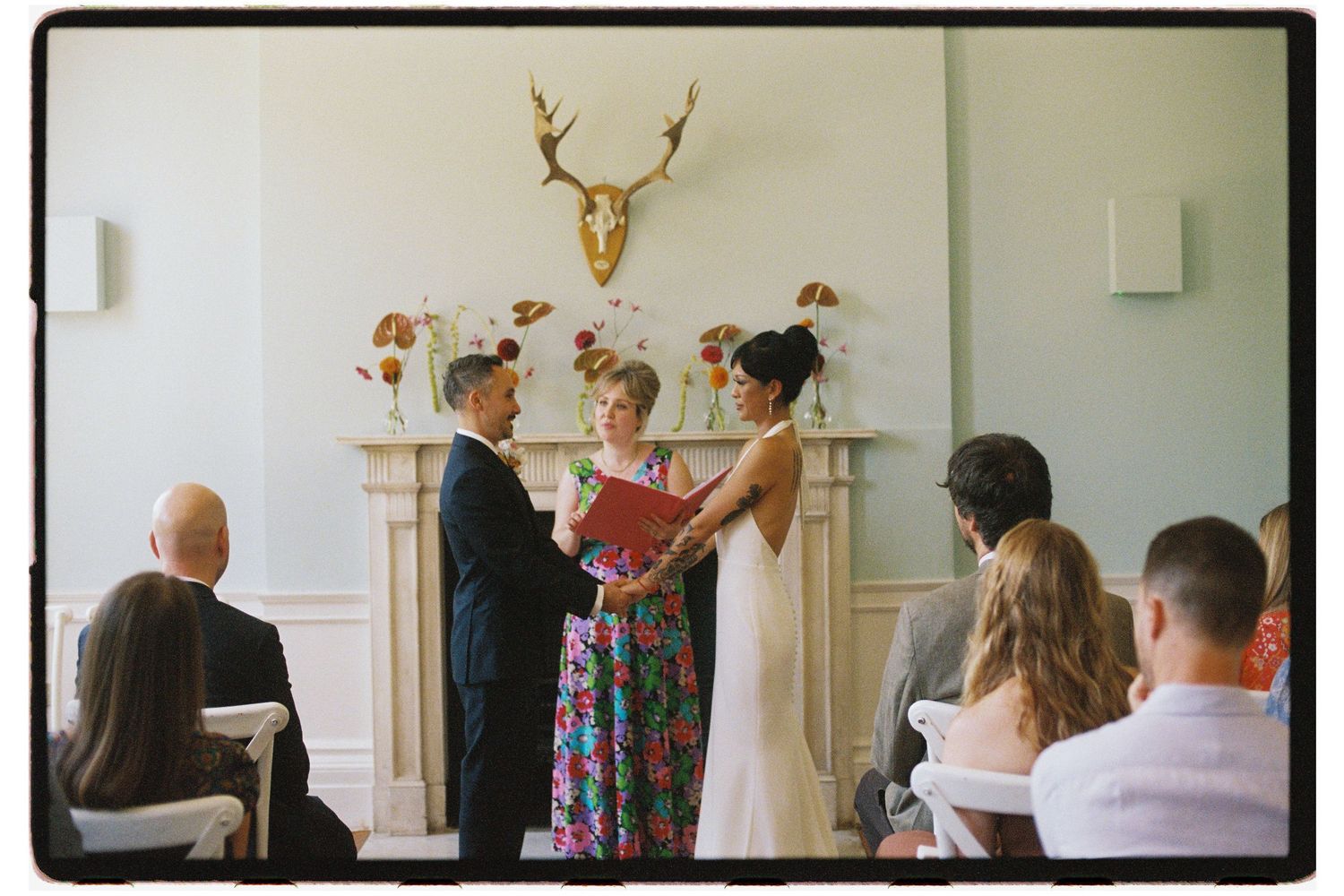 Couple exchanging vows at indoor wedding ceremony with officiant, floral arrangements, and gold deer head above fireplace.