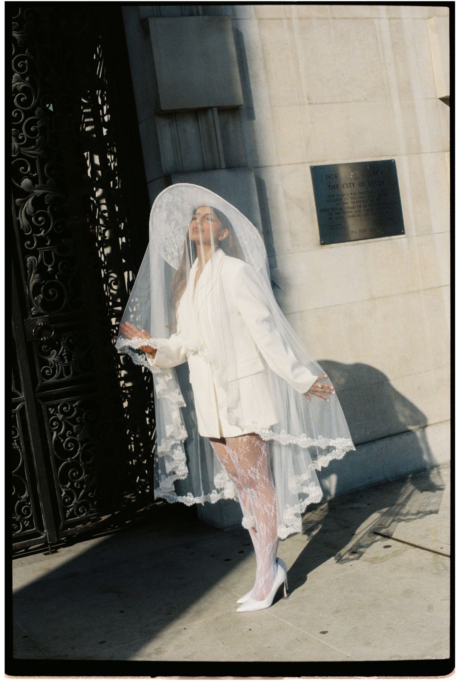 Fashion-forward bride in white blazer and lace veil posing dramatically at ornate iron gates.