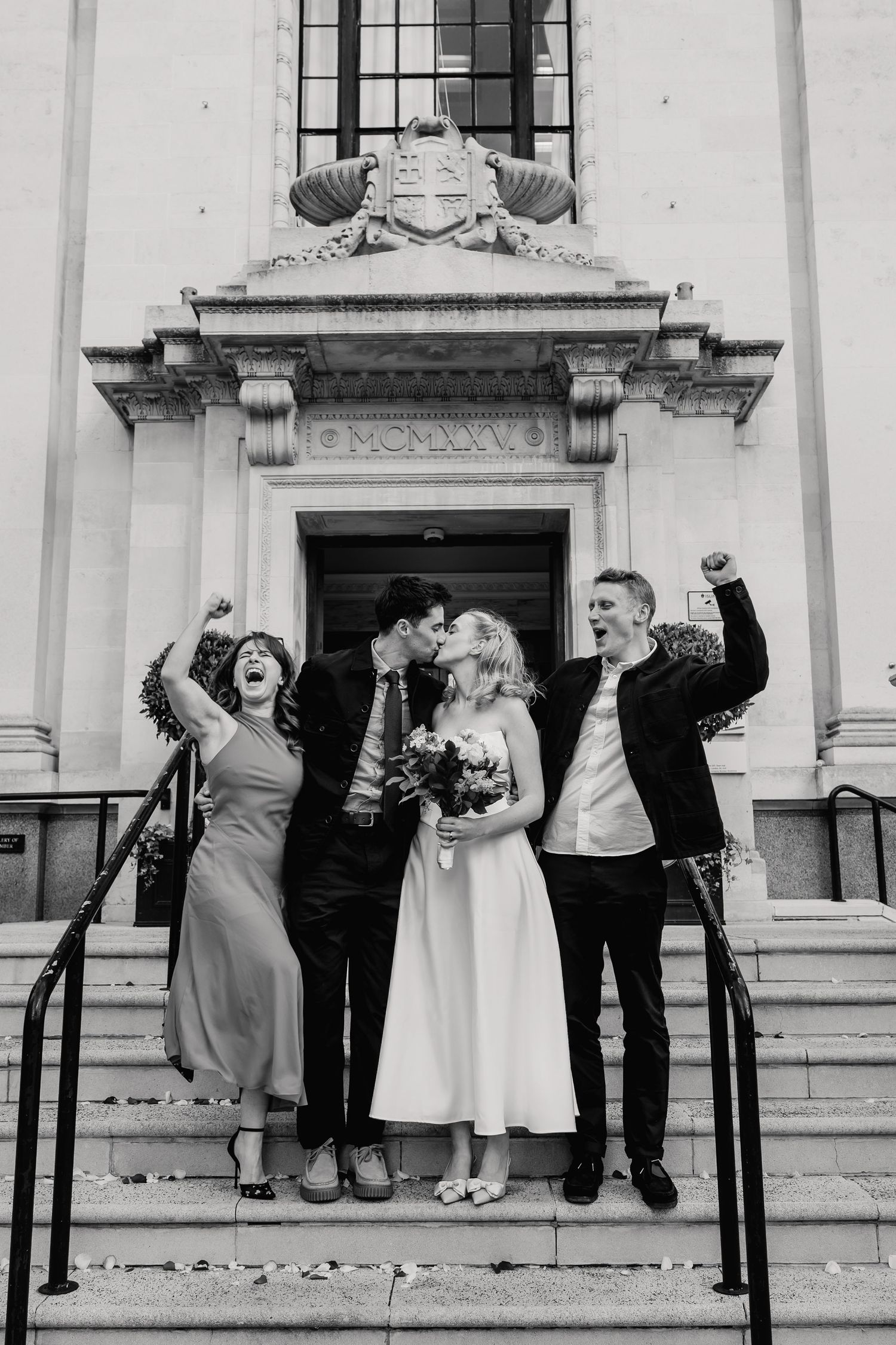 Newlyweds kissing on steps while two guests celebrate with raised fists outside registry office.