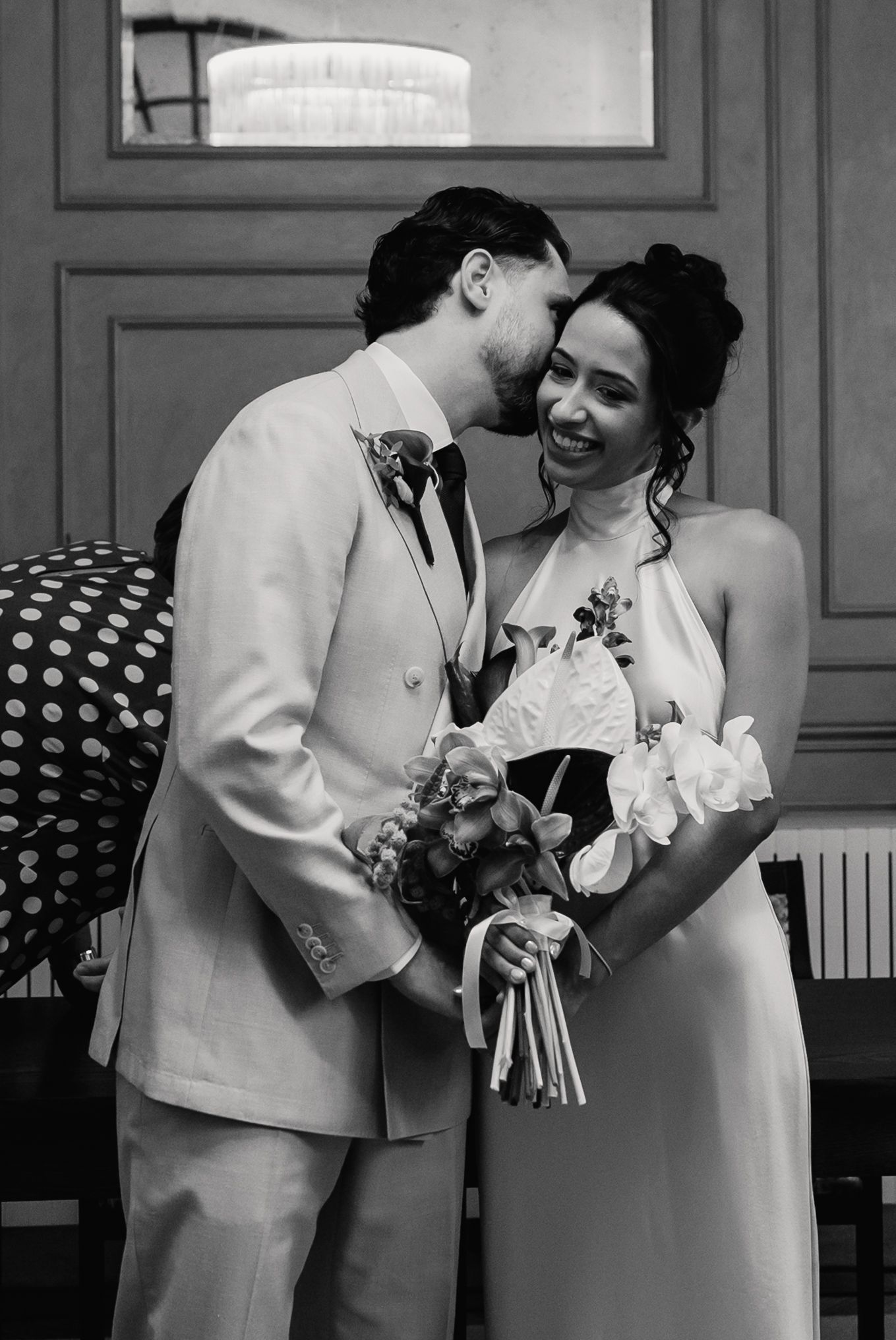 Black and white photo of groom in light suit kissing brides cheek as she laughs holding floral bouquet.