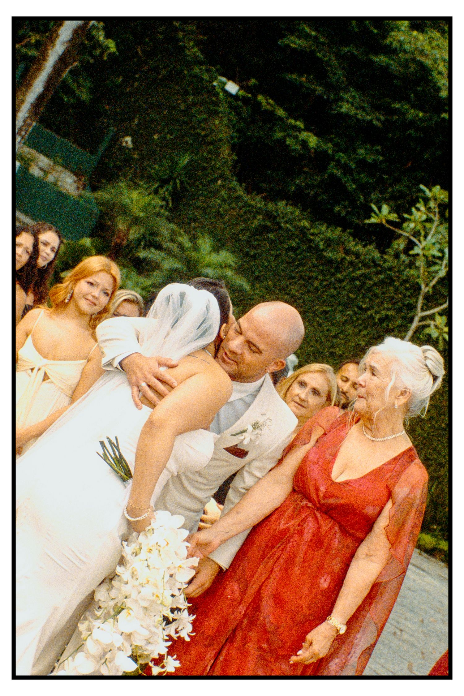 Groom lifting bride joyfully after ceremony surrounded by guests and lush green garden backdrop.