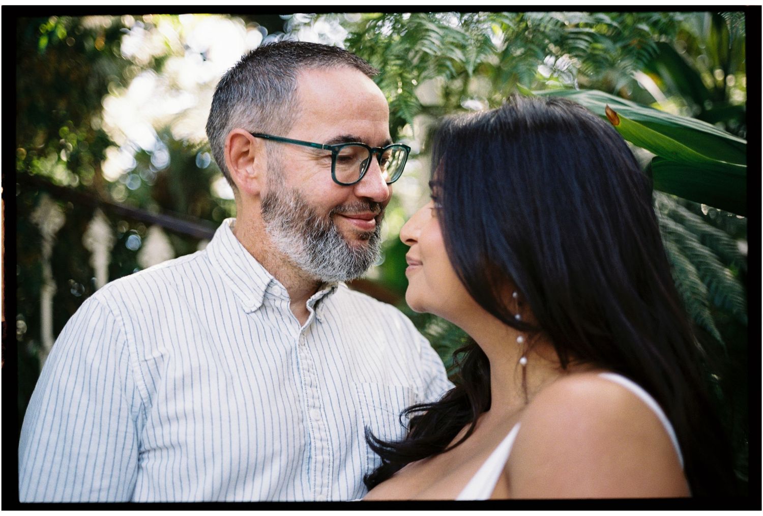 A couple shares a romantic moment outdoors, smiling at each other surrounded by lush green tropical foliage.