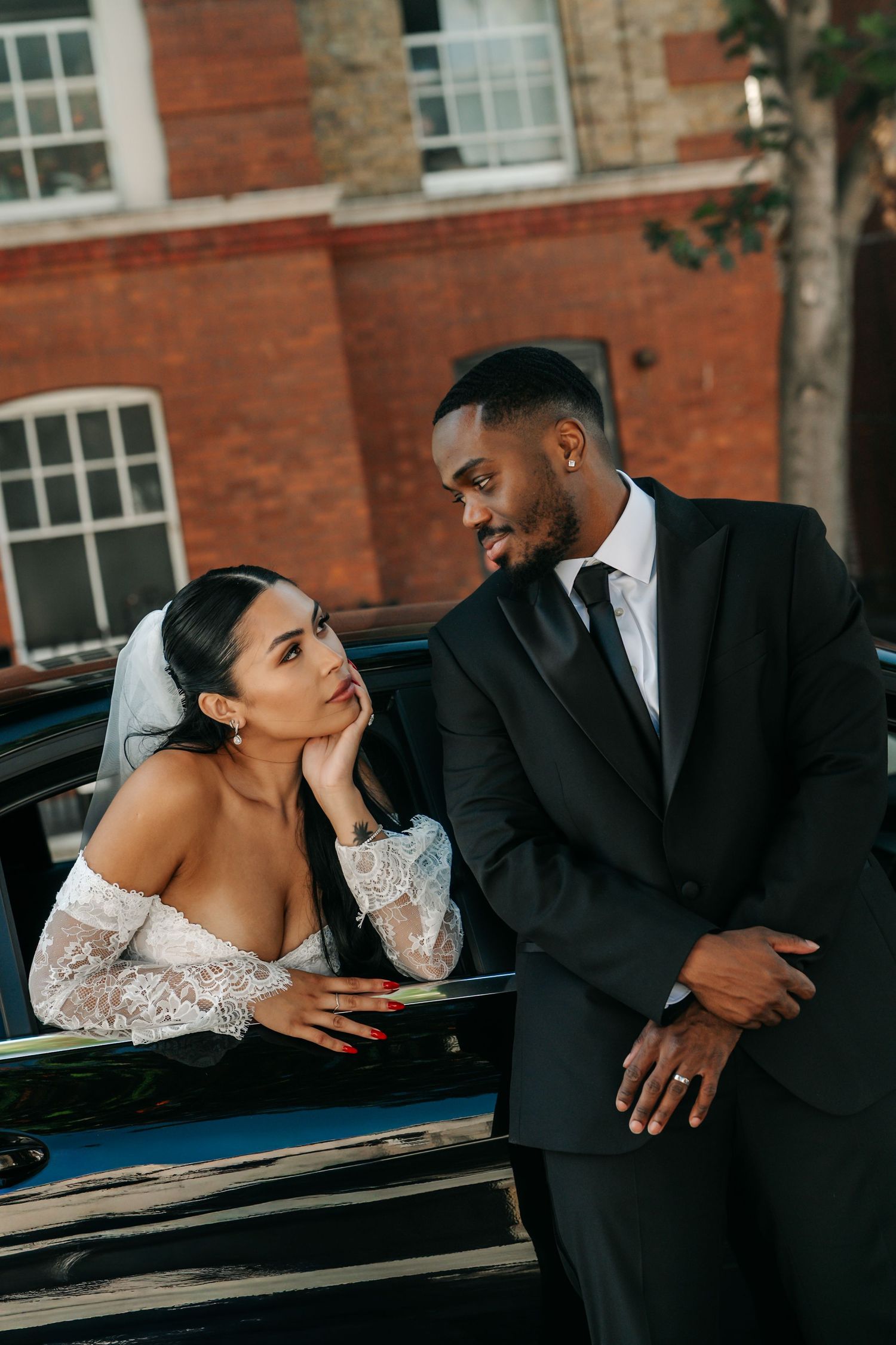 Bride in lace gown and groom in black suit share a romantic gaze beside a vintage car on their wedding day.