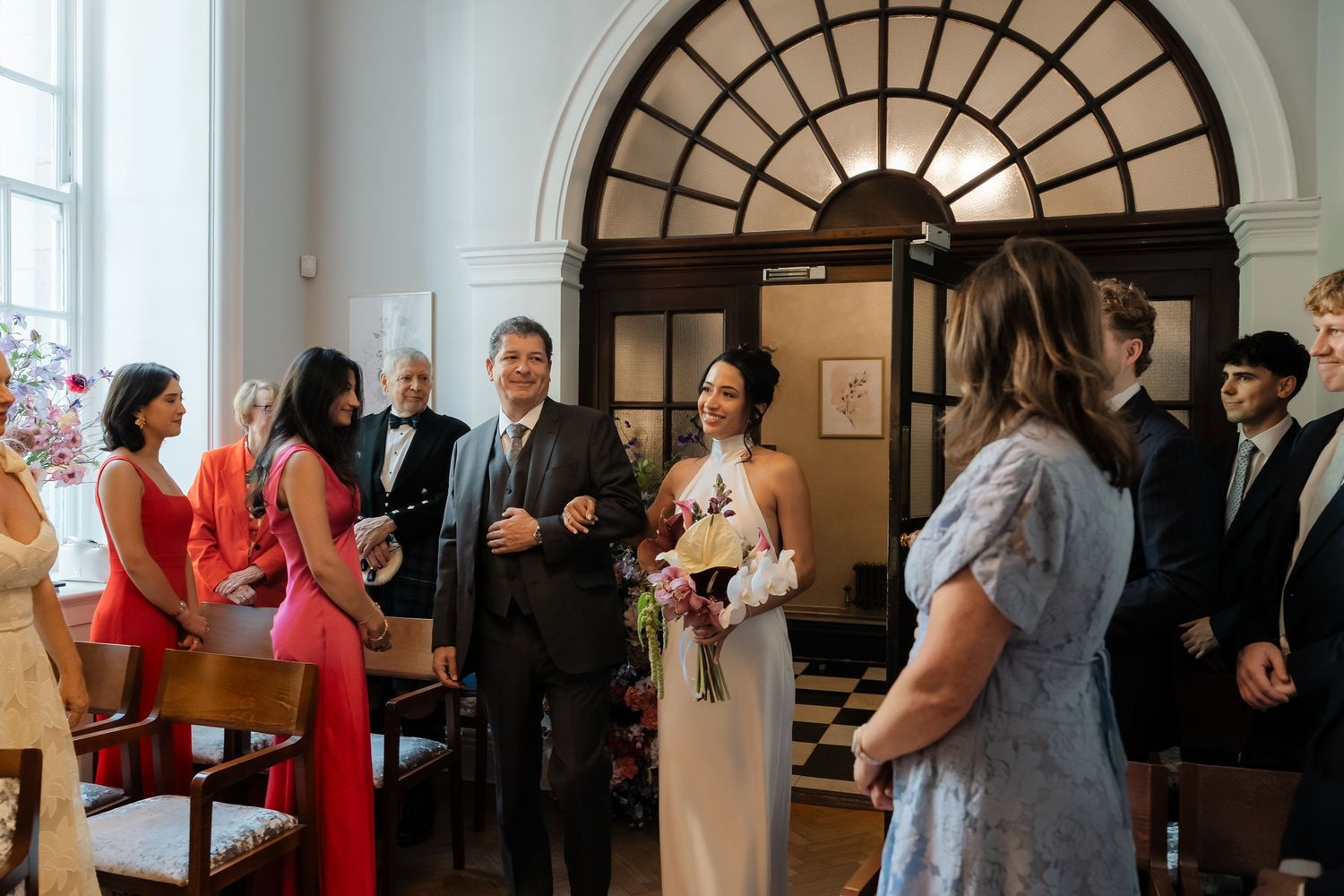 Bride holding a bouquet smiles at guests during an intimate wedding ceremony in an elegant venue.