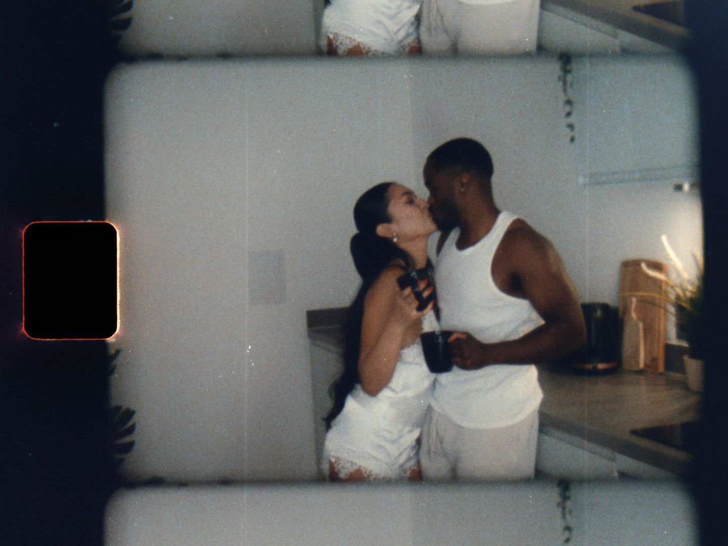 A couple shares a romantic kiss in a kitchen, both dressed in white outfits, captured in a vintage film photo style.