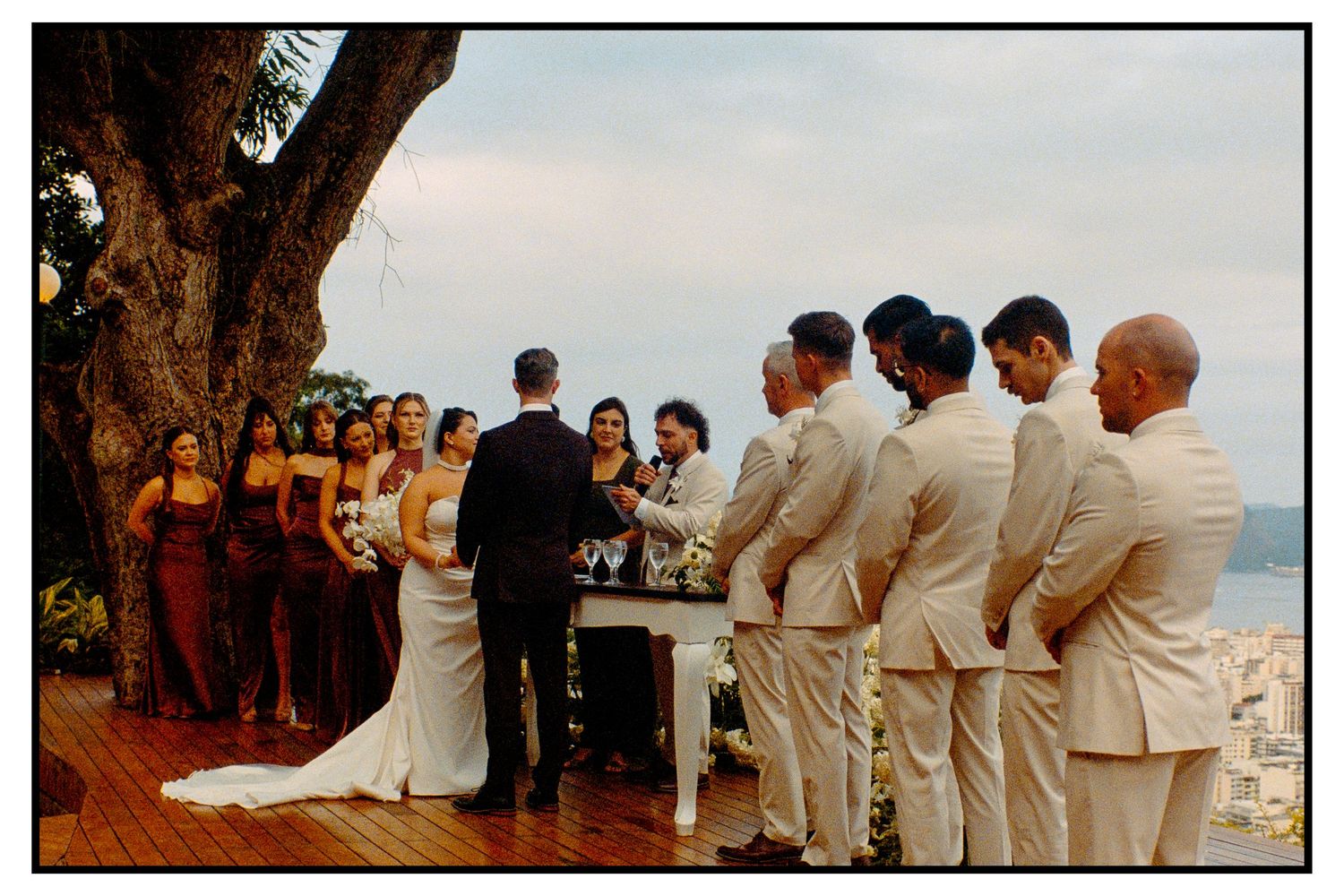 Outdoor wedding ceremony under large tree with bridesmaids in burgundy and groomsmen in beige by ocean.