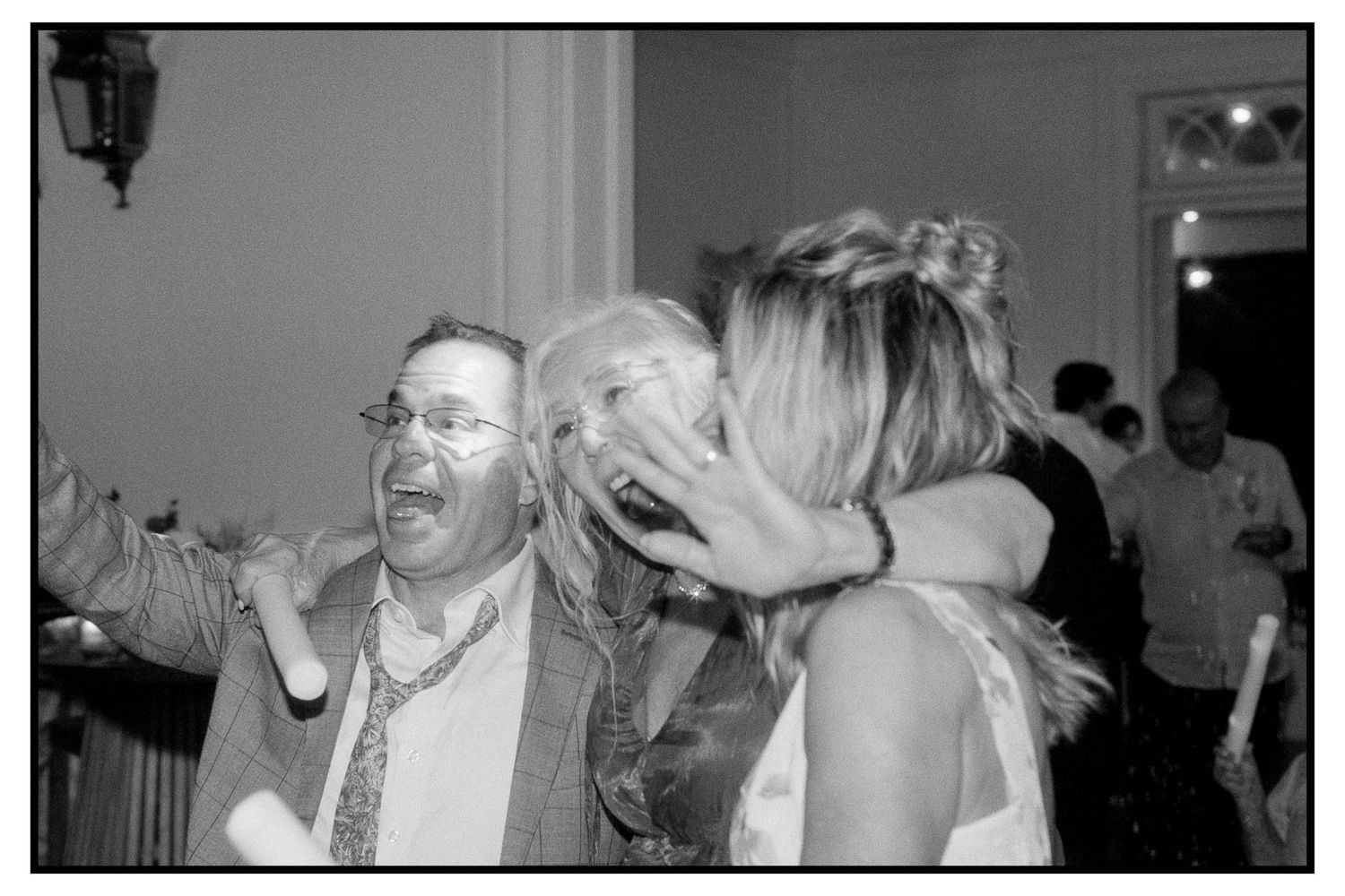 Black and white candid photo of wedding guests laughing and celebrating joyfully at reception.