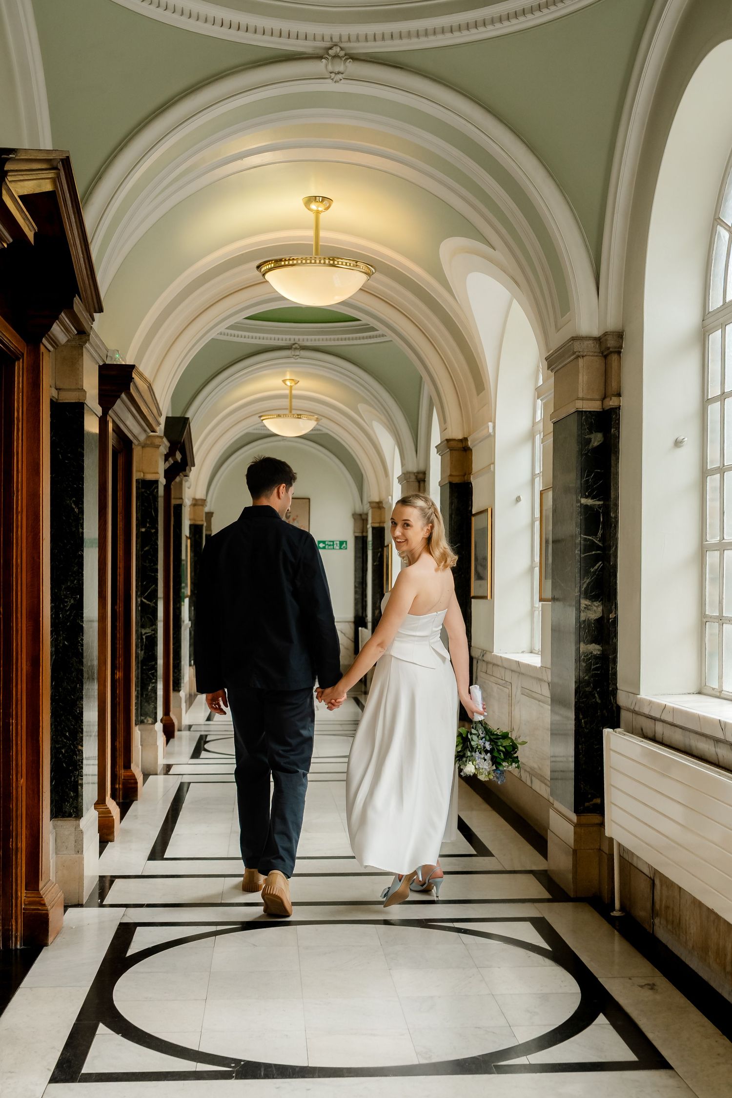 Newlywed couple holding hands walking through elegant arched marble hallway after civil wedding ceremony.
