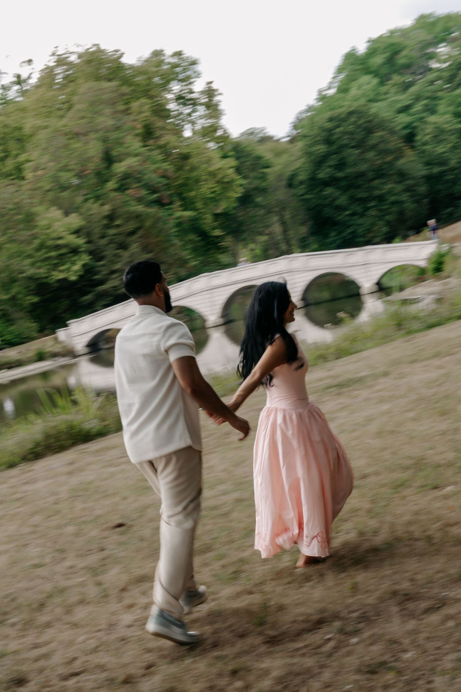 Couple holding hands walking through a park toward a stone arch bridge, woman wearing a flowing pink dress.