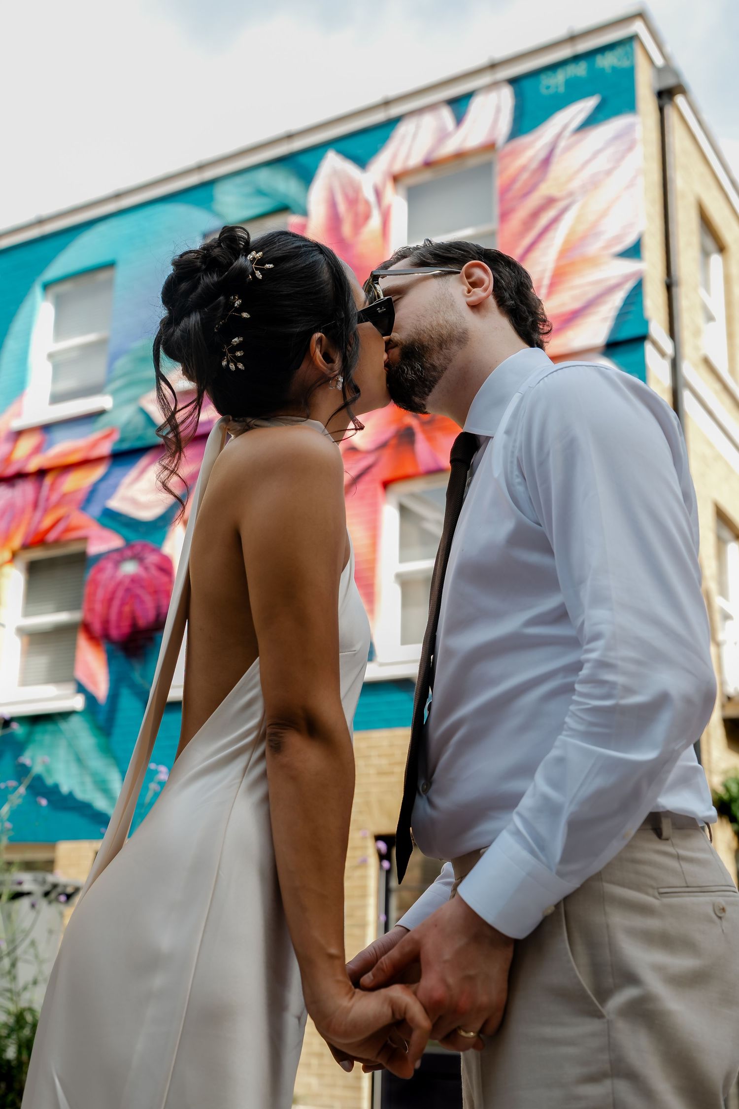 Bride and groom kissing while holding hands in front of colorful floral mural.