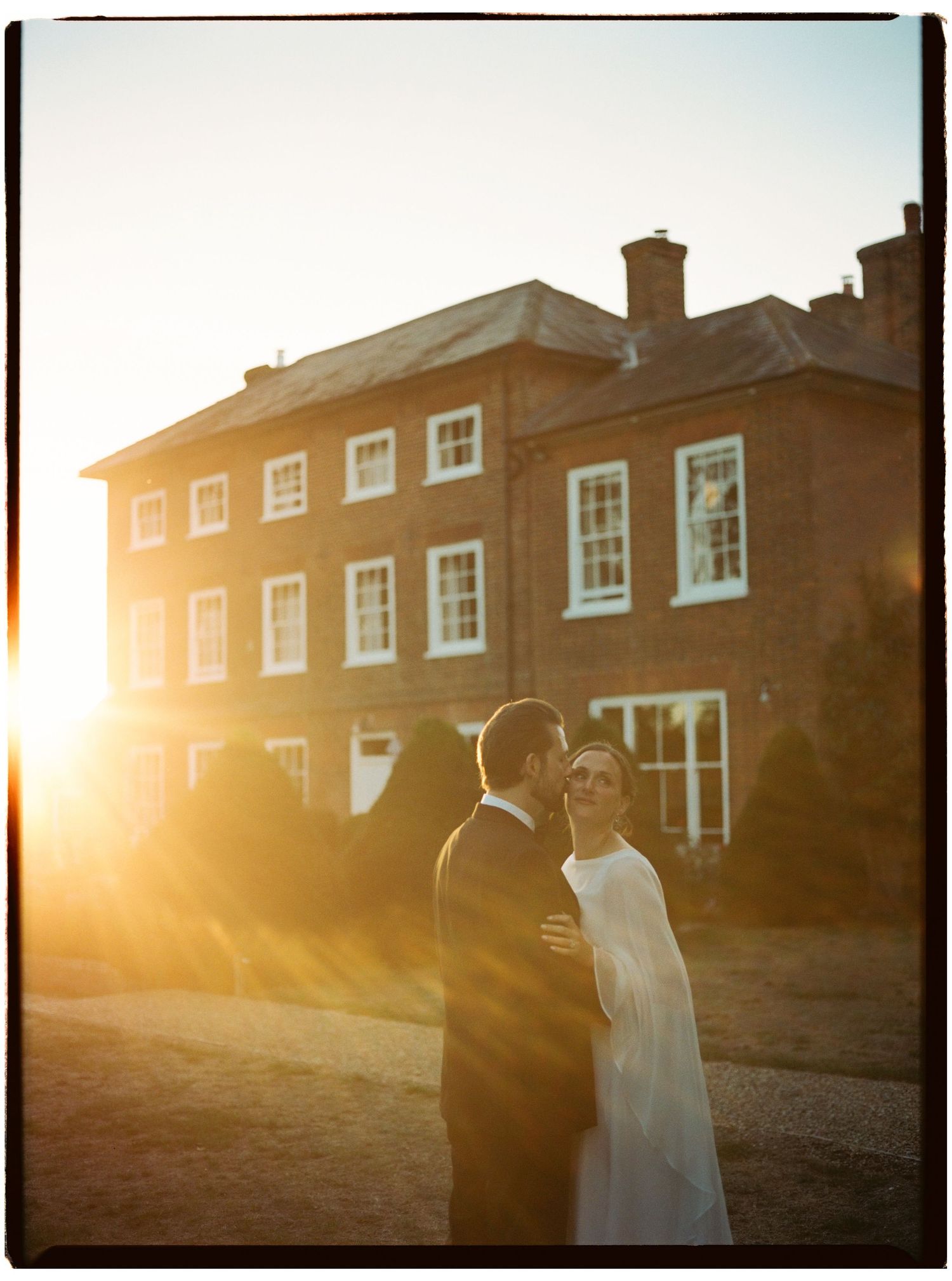 Couple embracing at golden hour sunset in front of a grand red brick manor house on their wedding day.