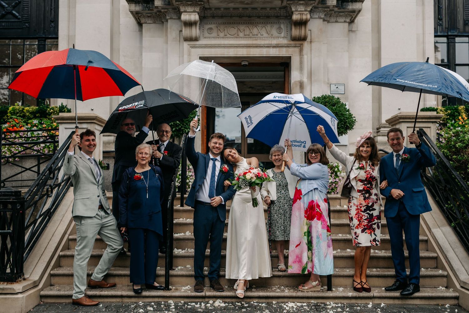 Wedding party celebrating on rainy steps, holding colorful umbrellas, bride in white gown with bouquet, guests in formal attire.