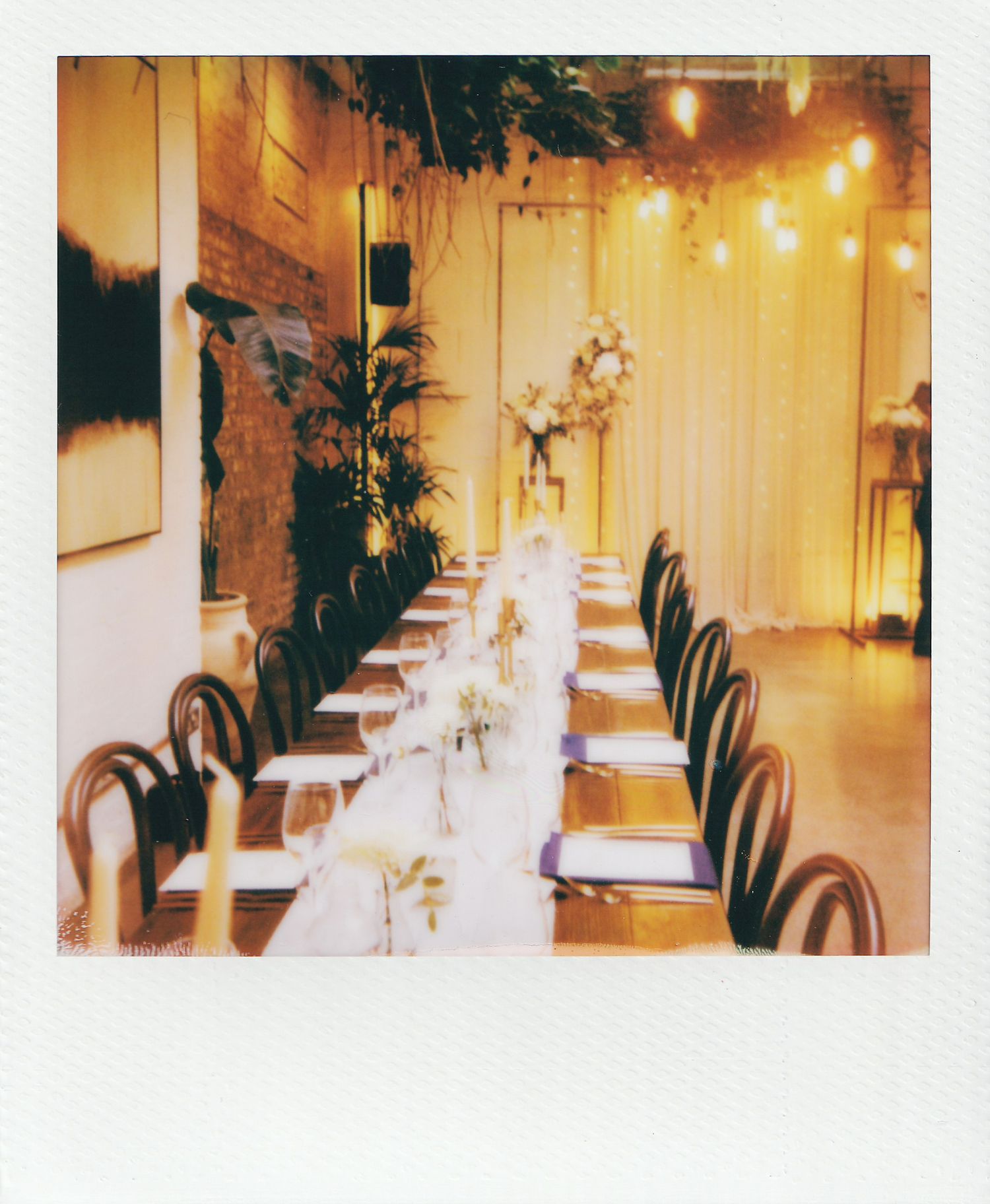 Elegant long dining table set for an event with string lights, white linens, and lush greenery in a warm, intimate venue.