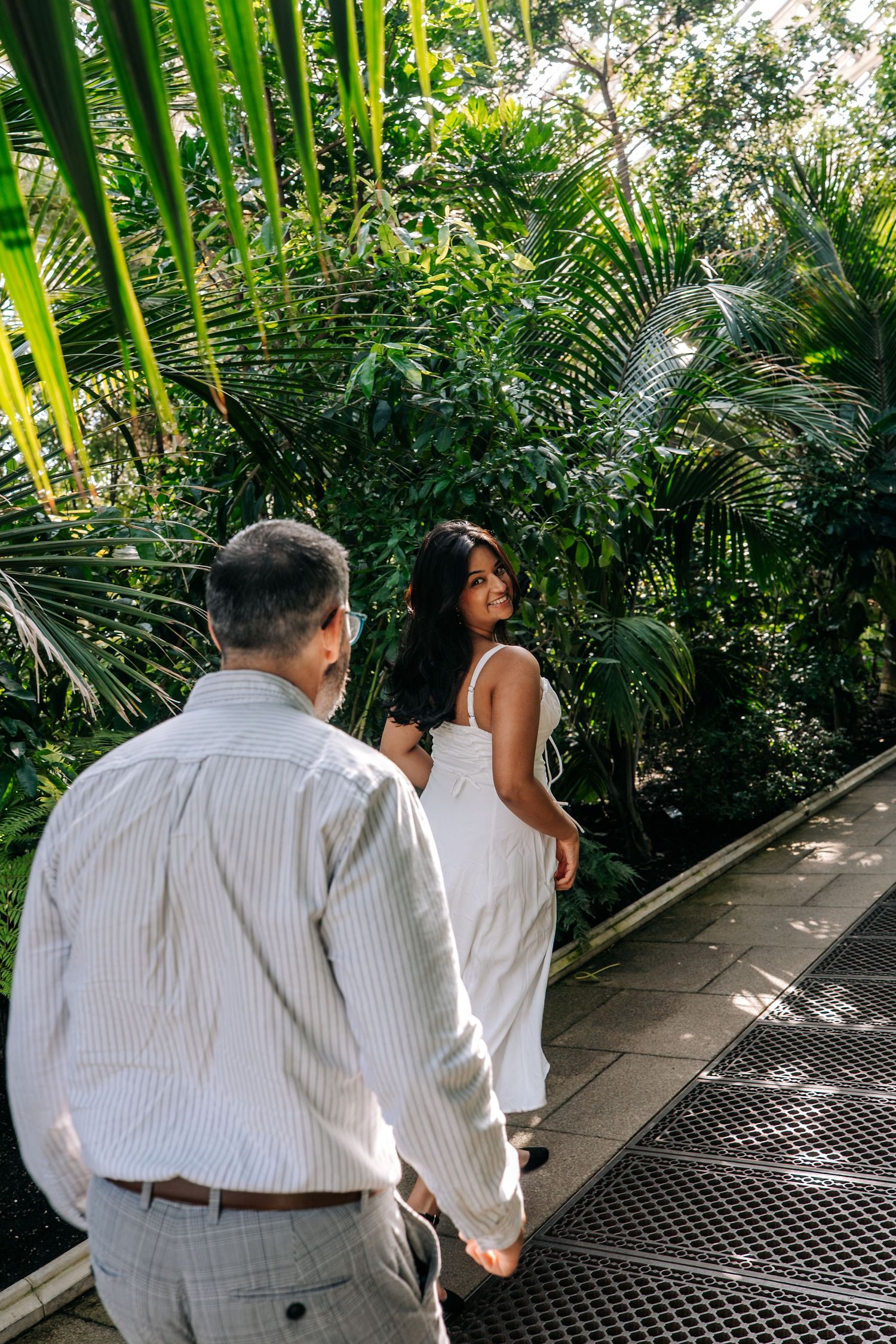 Couple walking hand-in-hand through a lush tropical garden, woman in white dress glancing back.