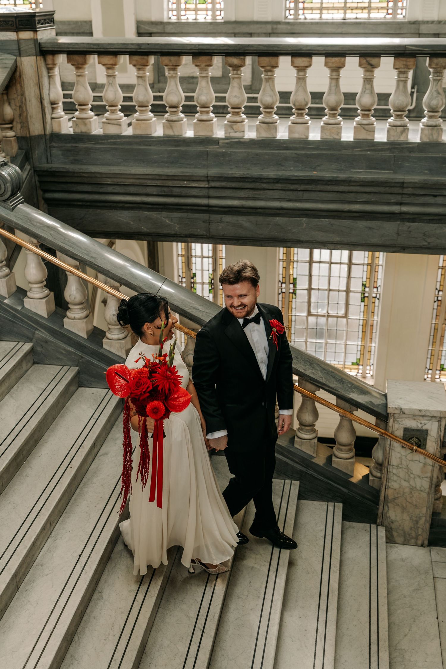 Bride and groom walking down grand marble staircase, bride holding red floral bouquet, groom in black tuxedo.