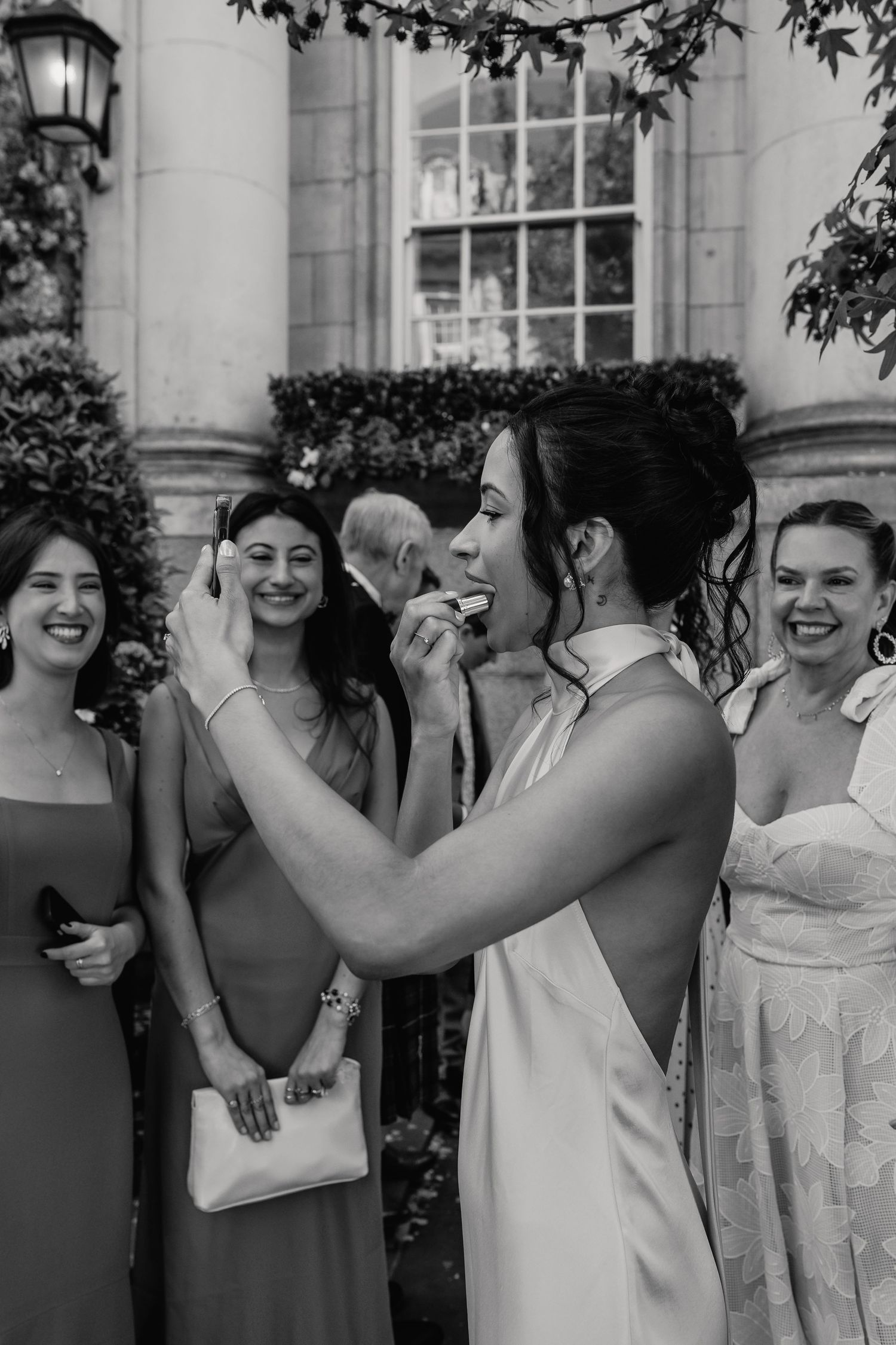 Bride in white halter dress adjusting earring with guests laughing behind her.