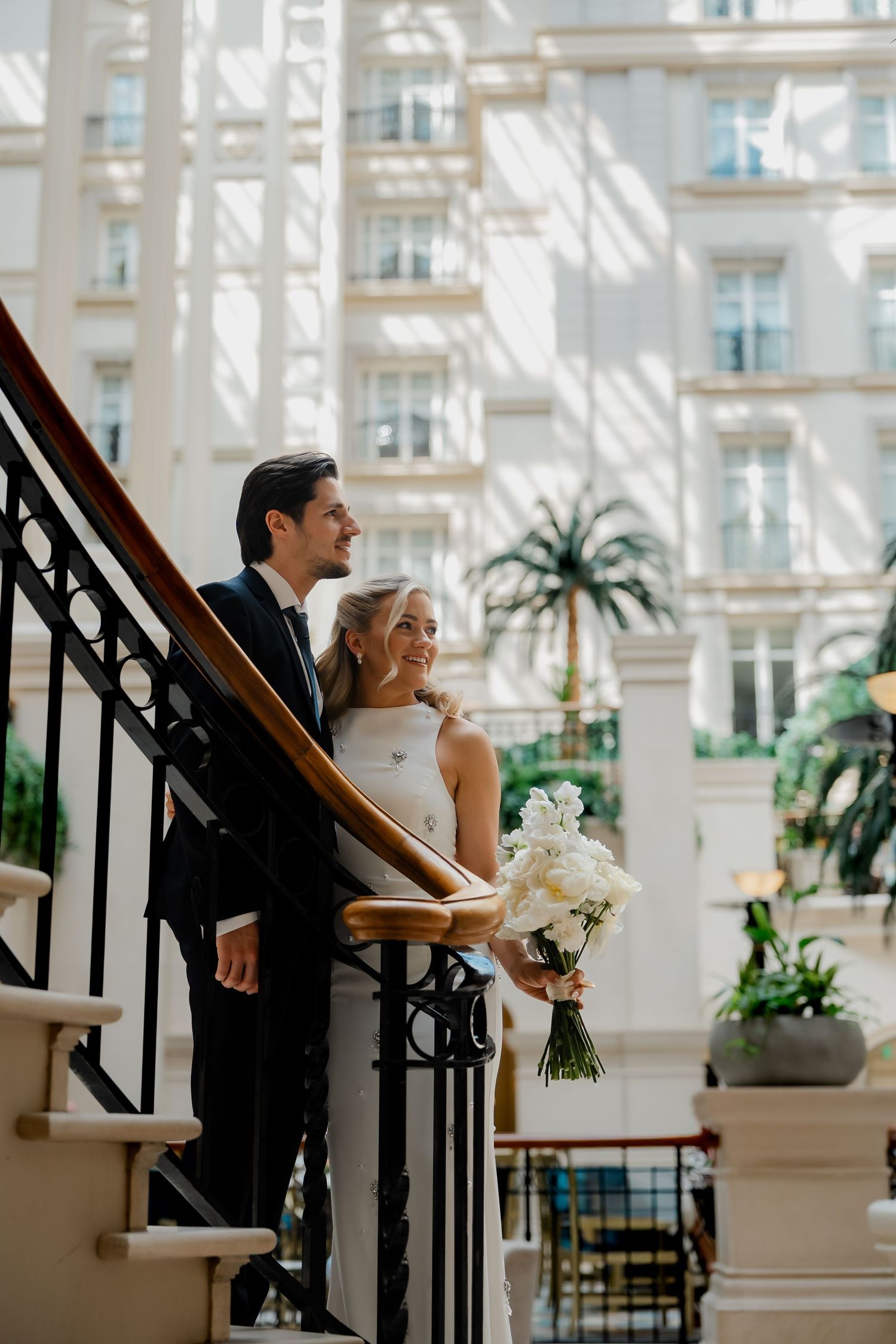 Happy bride and groom standing on a sunlit grand staircase with the bride holding a white bouquet.
