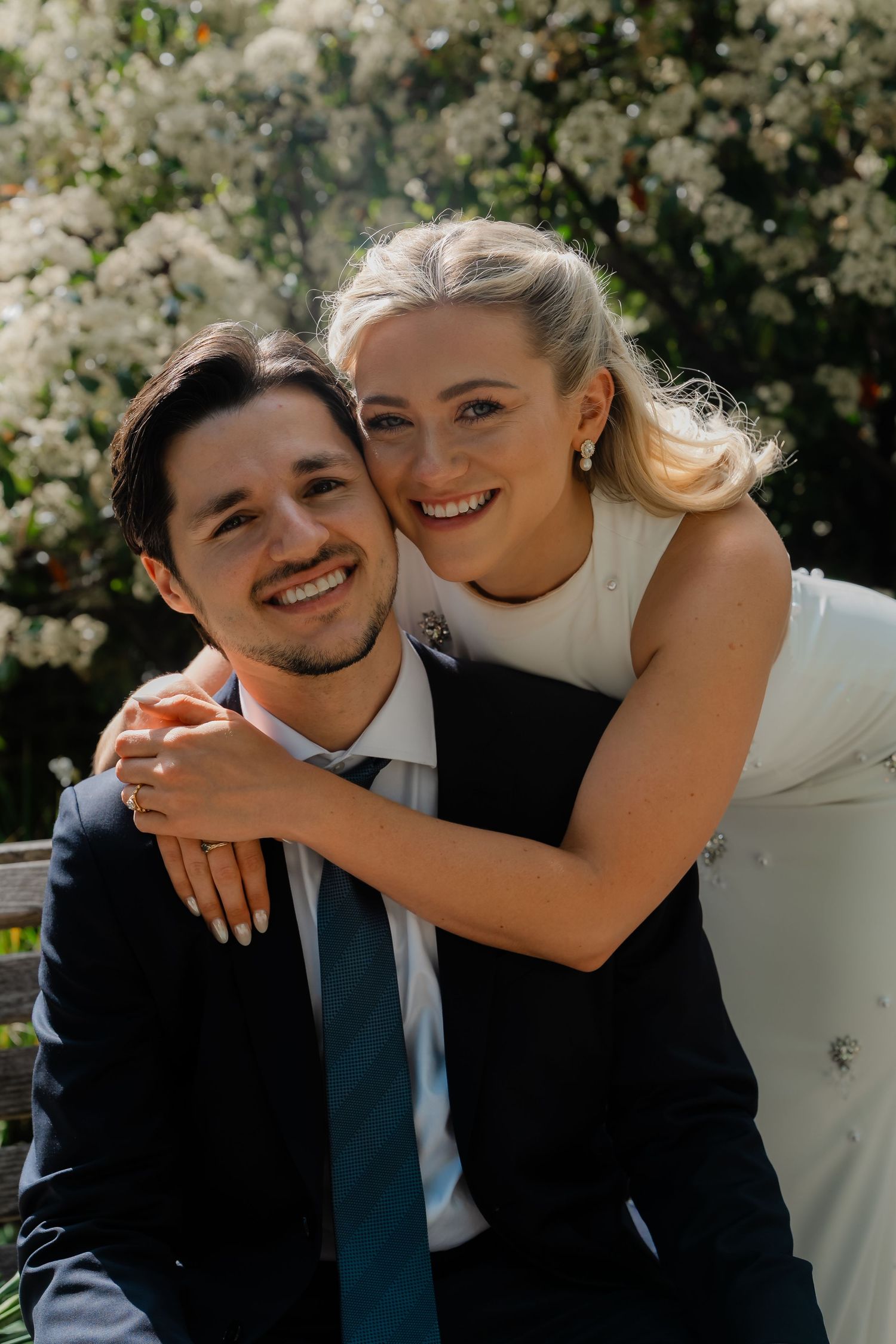 Smiling bride hugs seated groom on a bench surrounded by blooming white flowers outdoors.