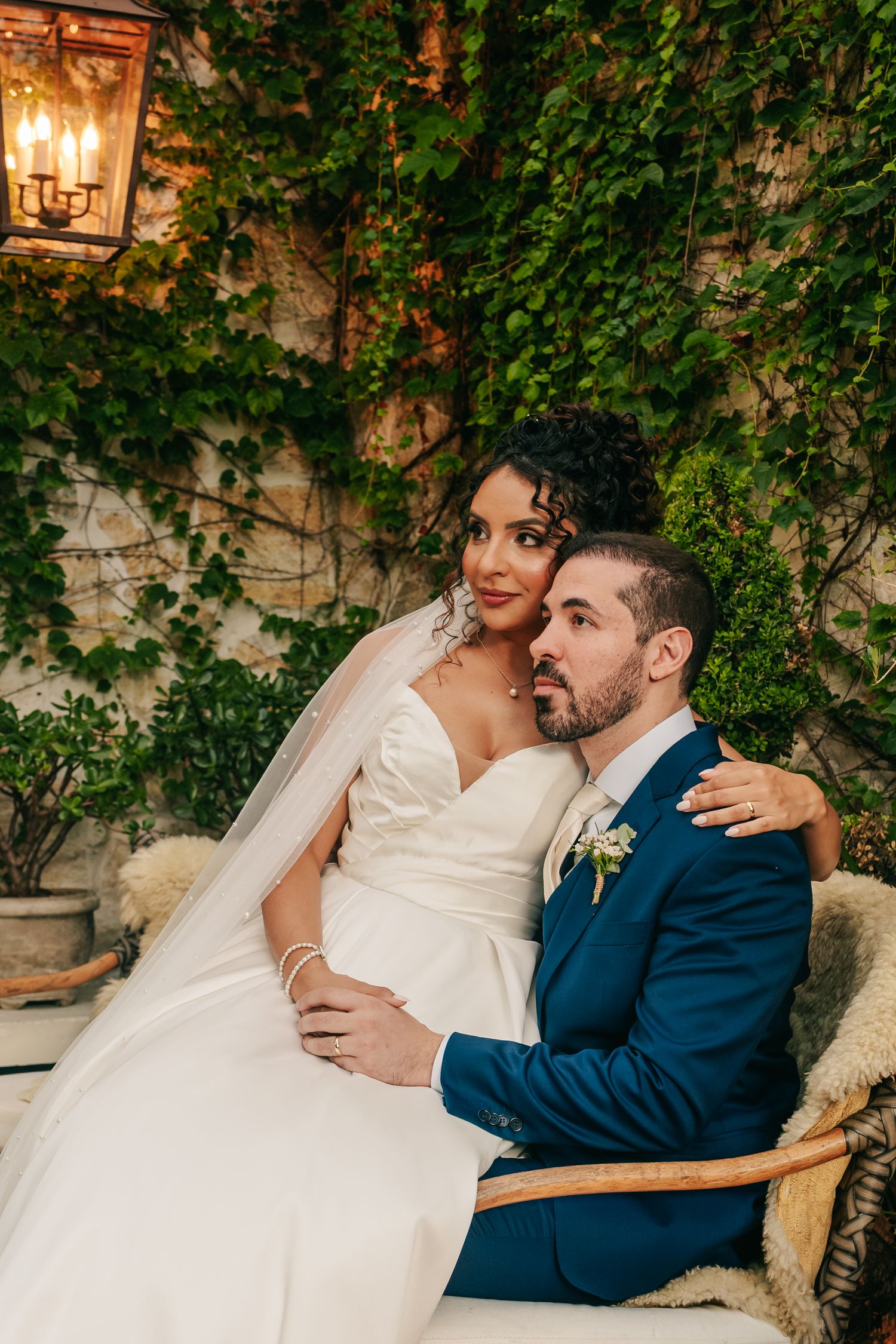 Bride in pearl veil sits with groom in navy suit against romantic ivy-covered backdrop.