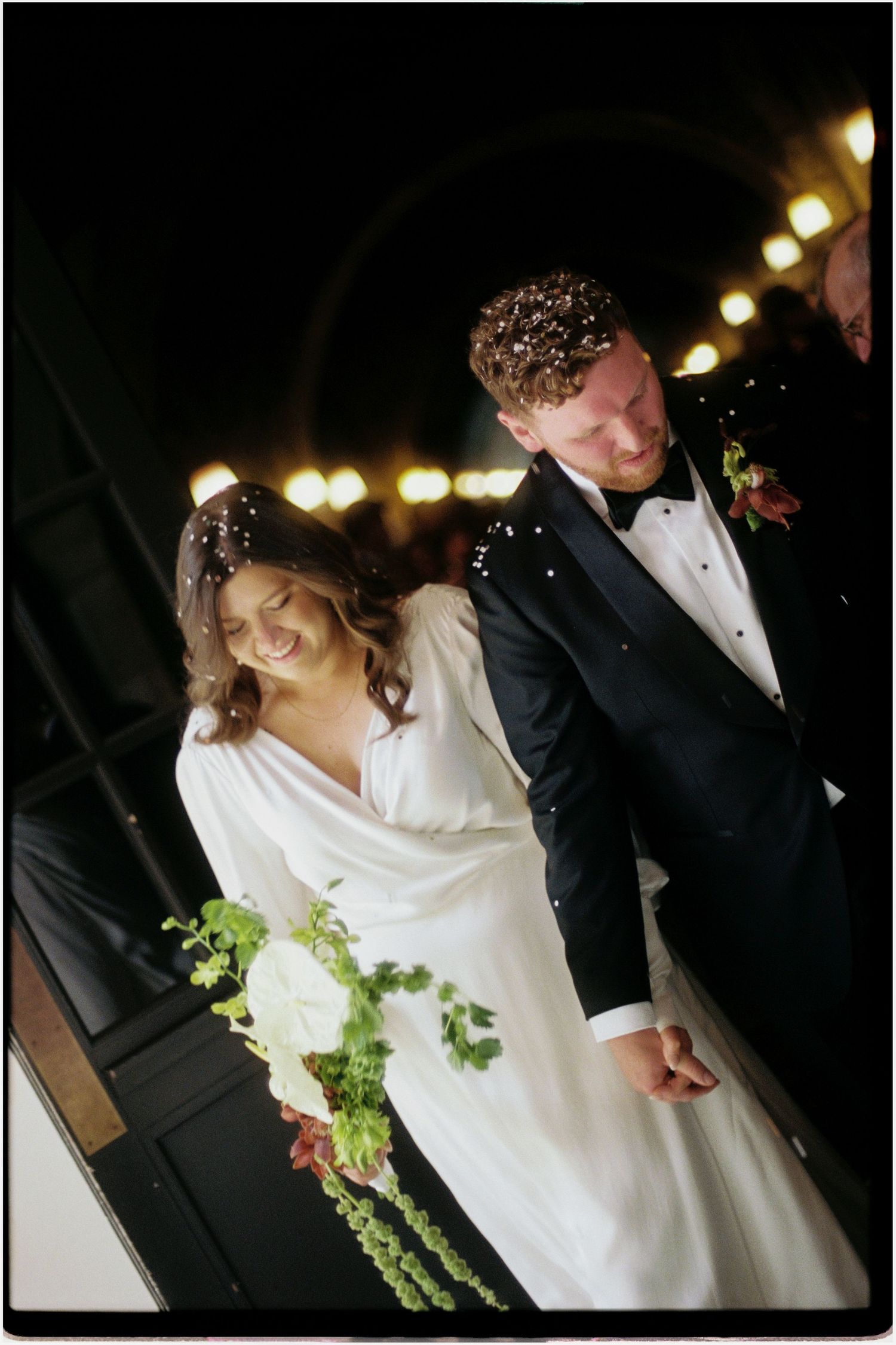 Bride and groom walking hand in hand under string lights at a nighttime wedding celebration.