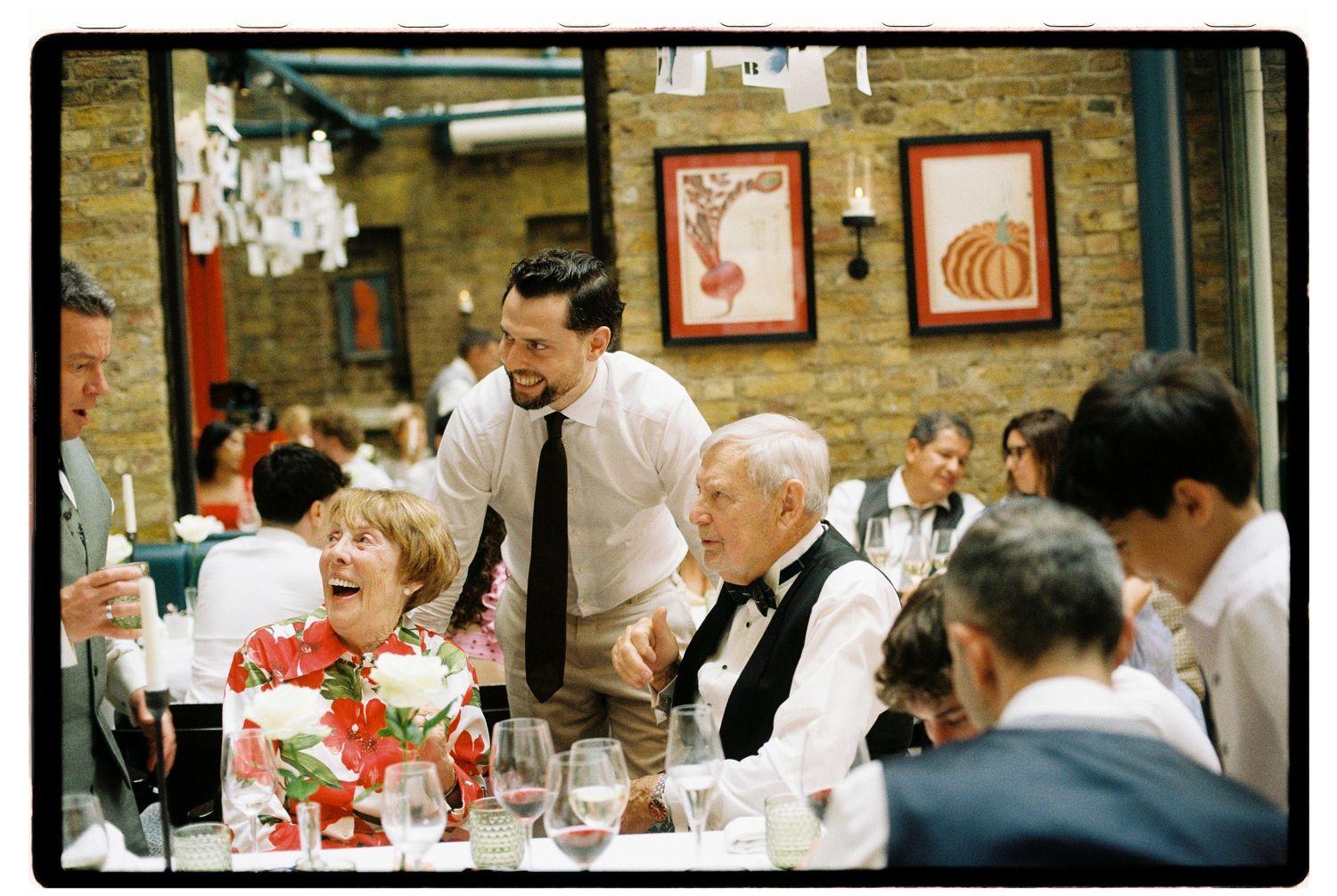 Groom in white shirt and tie laughing with elderly guests at colorful wedding reception dinner table.