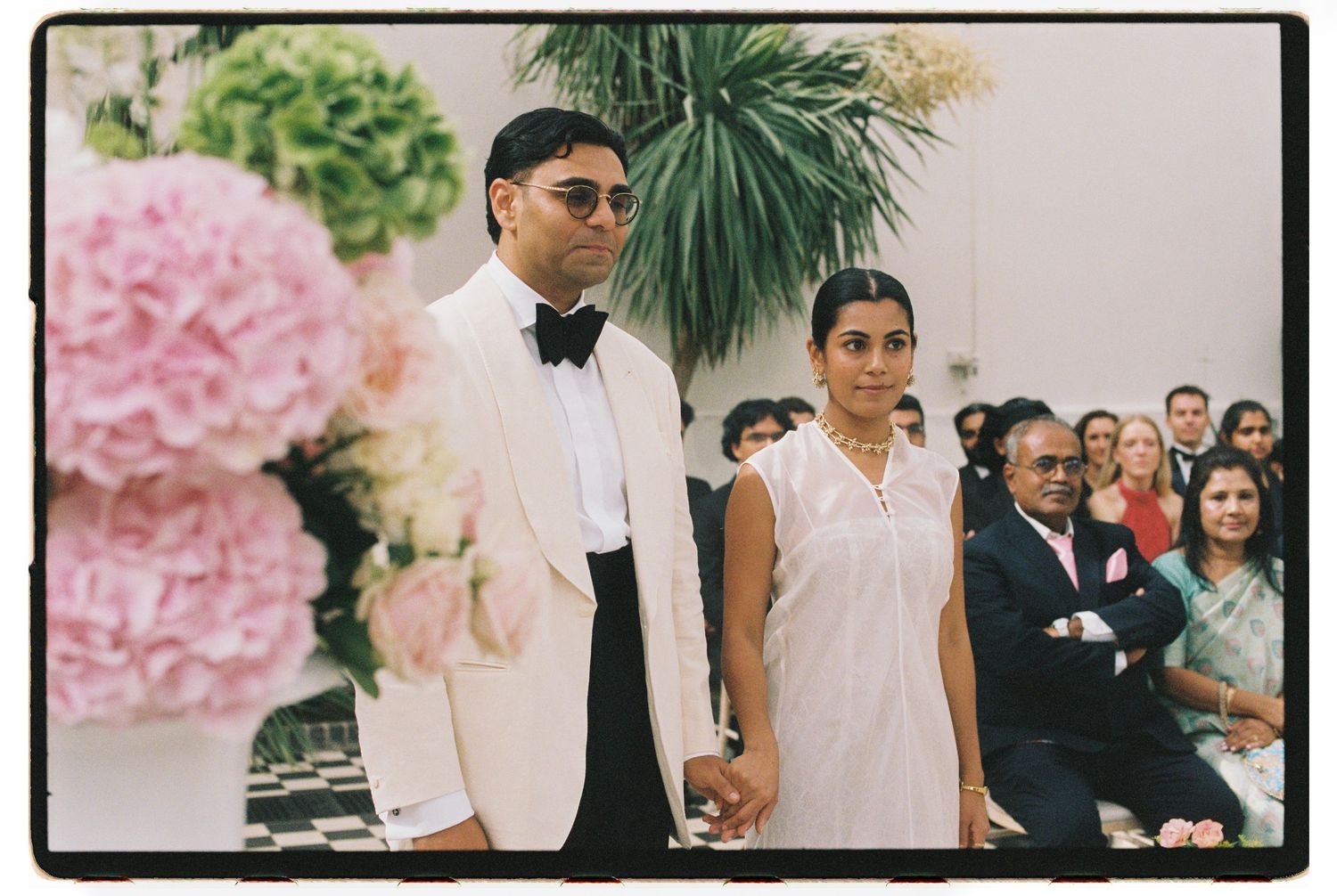 Couple in wedding attire holding hands at aisle, pink floral arrangements, guests seated in bright venue.