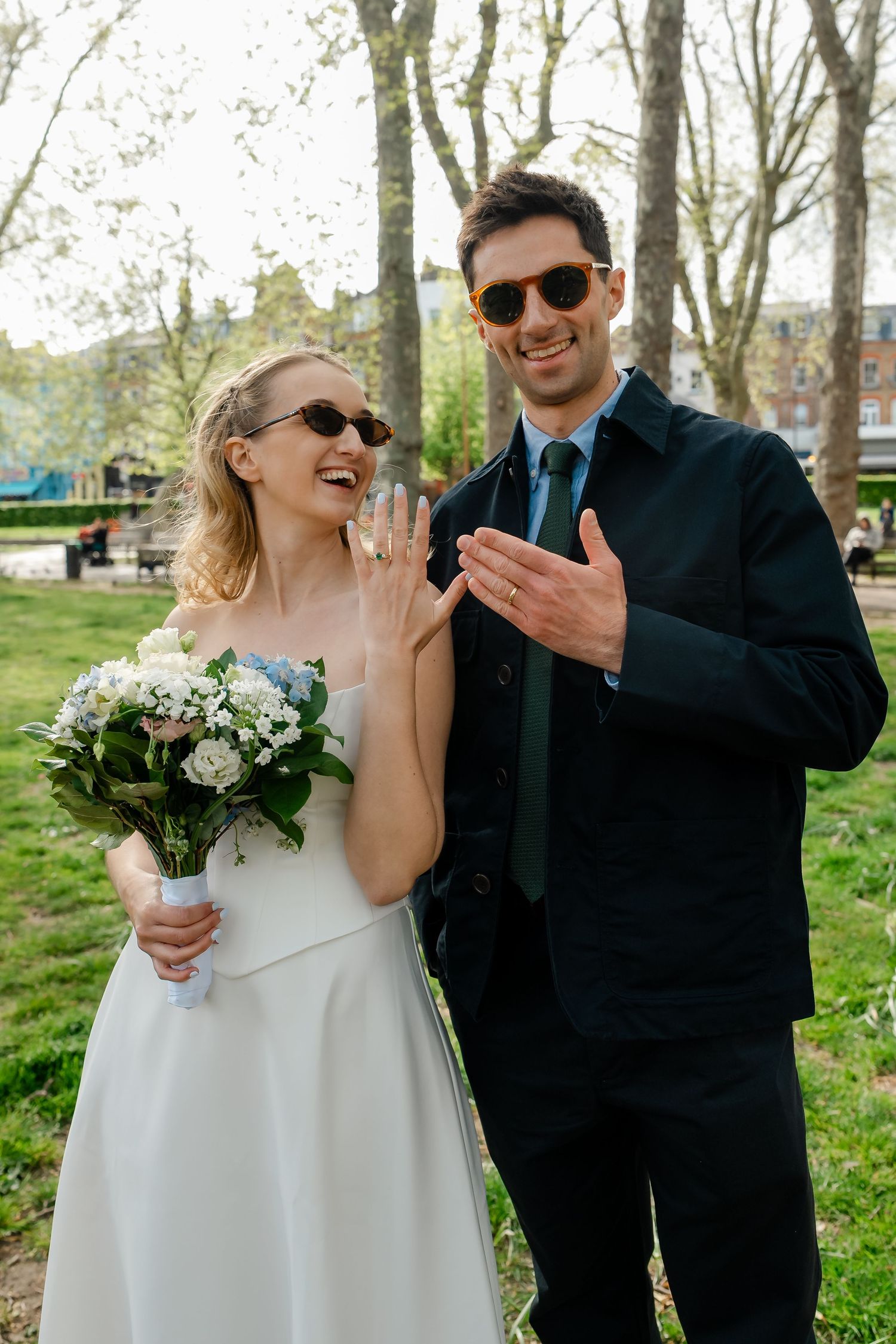 Newlyweds in sunglasses showing wedding ring while bride holds white bouquet in a sunny park.