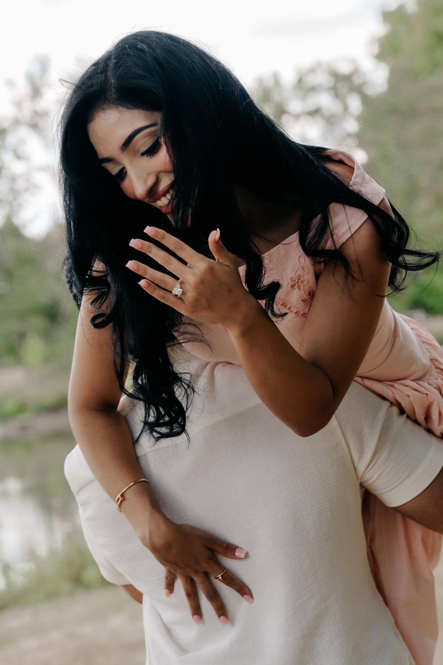 Joyful woman showing engagement ring while being lifted by partner outdoors after proposal.
