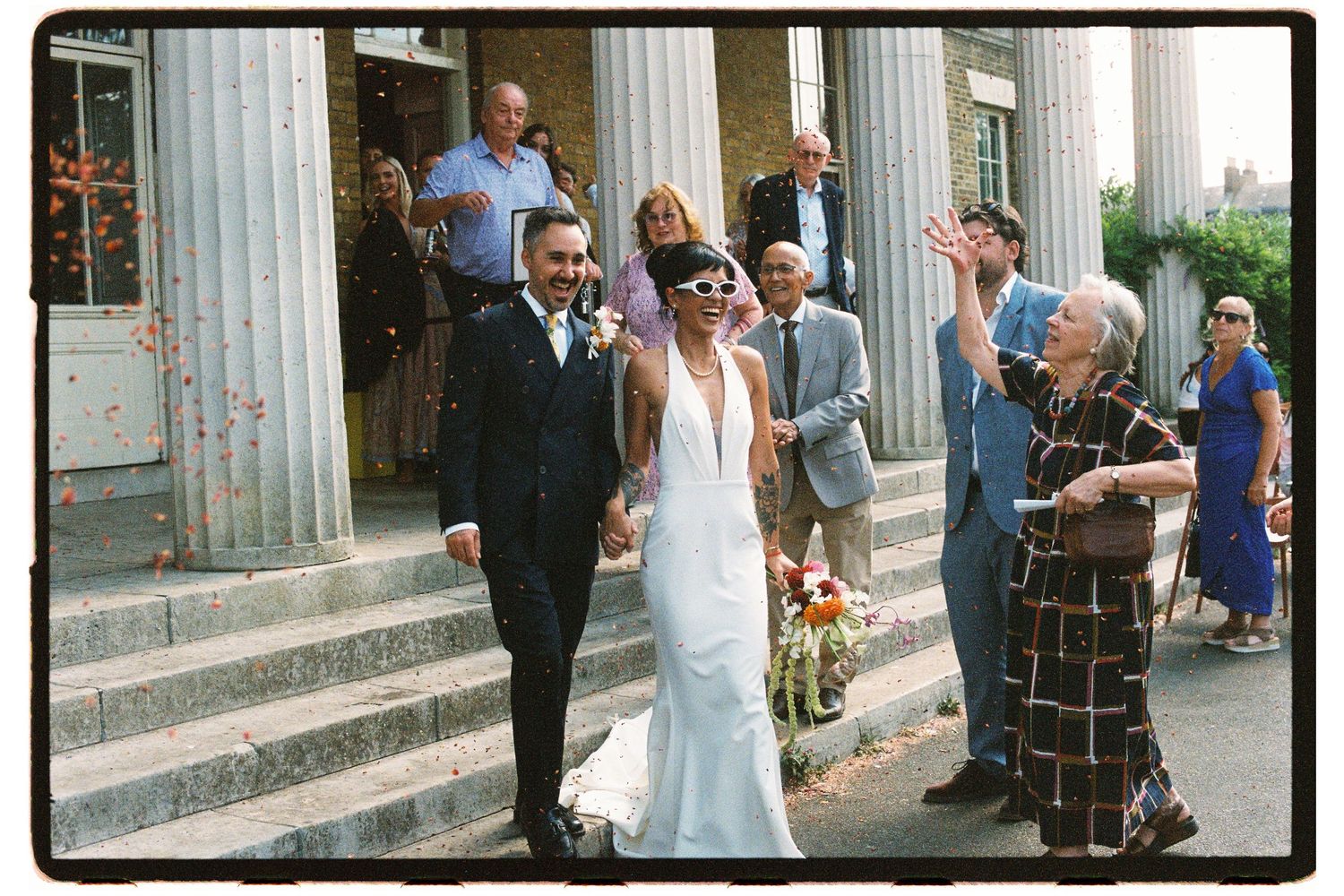Smiling newlyweds exit a grand columned building as guests celebrate with confetti.