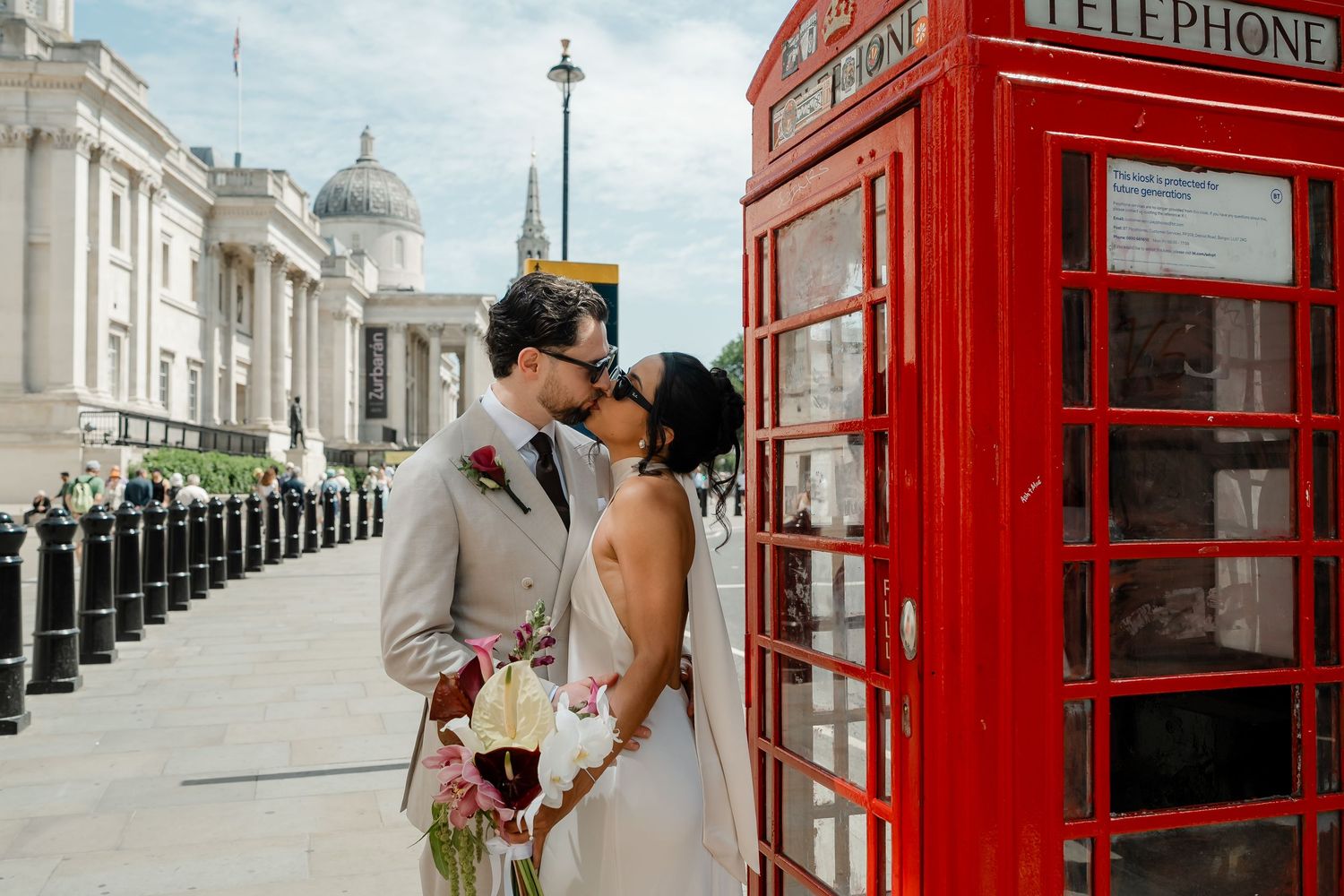 Bride and groom kissing beside iconic red London telephone box near Trafalgar Square on their wedding day.