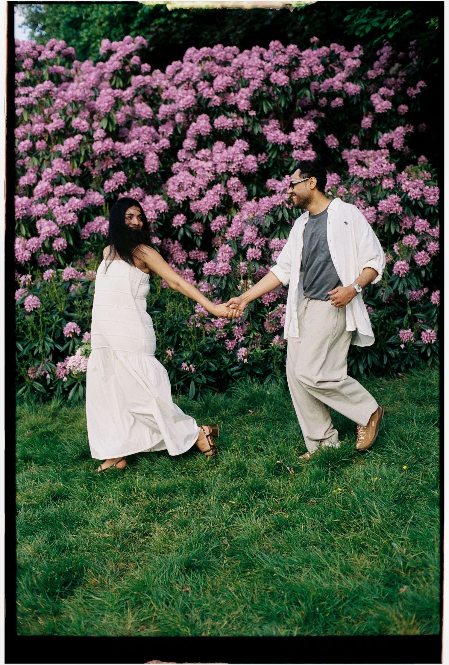 Couple holding hands dancing in lush garden with vibrant pink rhododendron bushes in bloom.