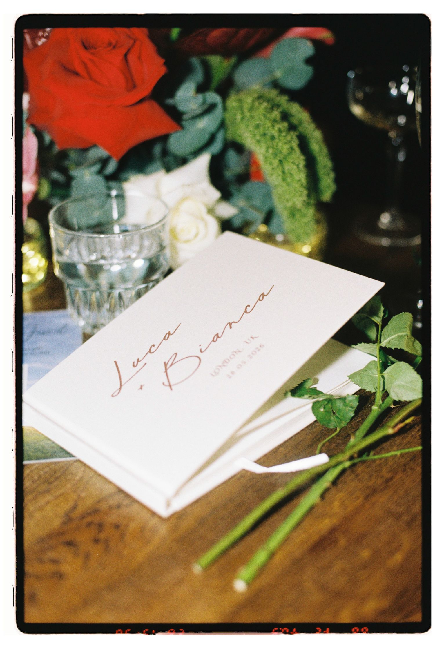 White wedding program booklet for Luca and Bianca on a wooden table with red roses and a glass.