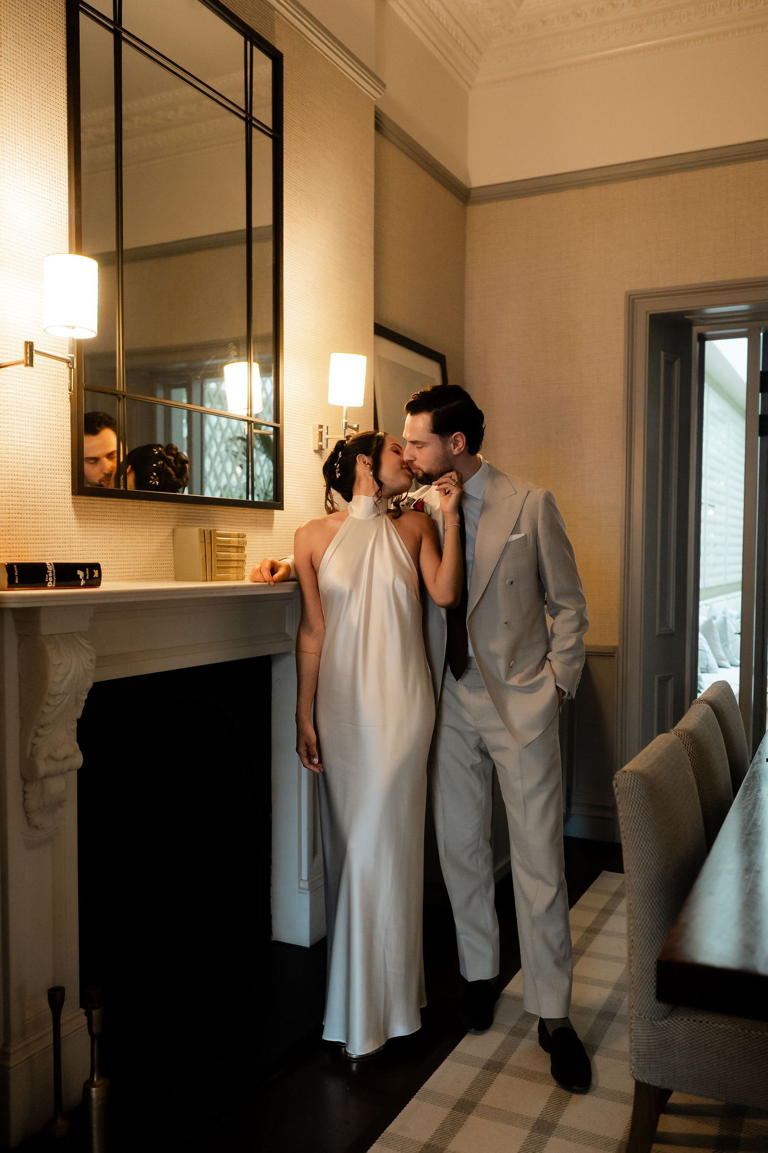 Bride and groom sharing an intimate moment by a fireplace, she in a silk halter gown, he in a beige suit.