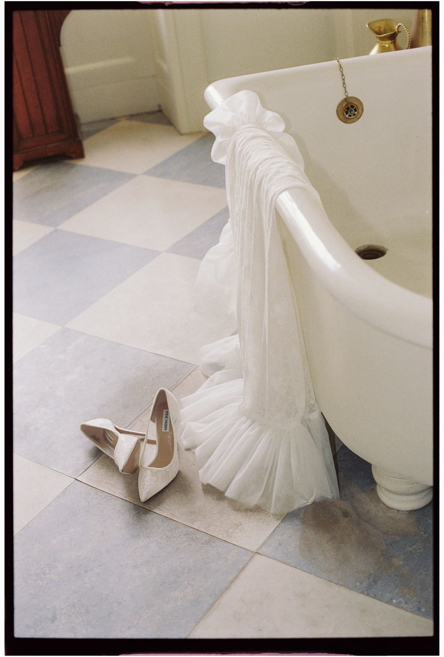 White wedding veil draped over a vintage clawfoot bathtub beside bridal heels on checkered floor.