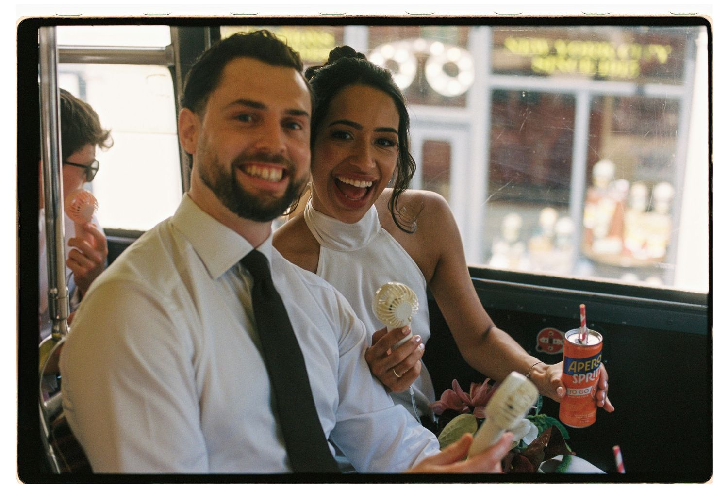 Smiling bride and groom seated inside a vintage bus, bride holding ice cream cone and colorful drink, color photo.
