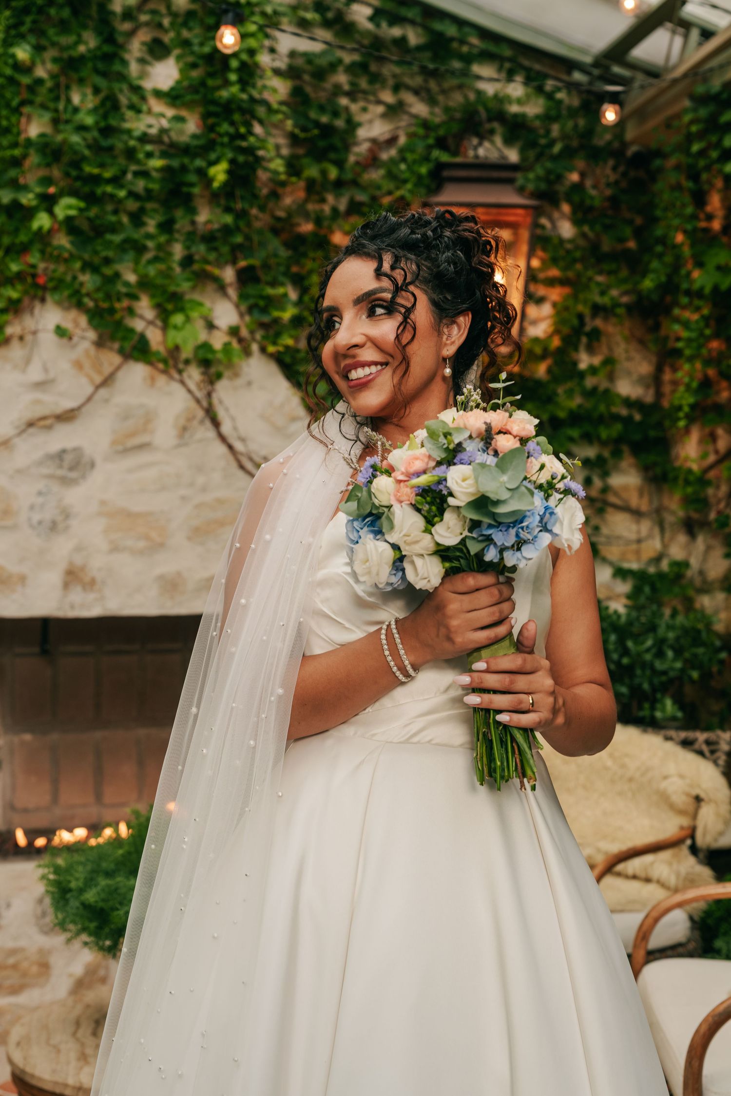 Smiling bride in white gown and pearl veil holds blue and pink bouquet before ivy-covered stone fireplace.