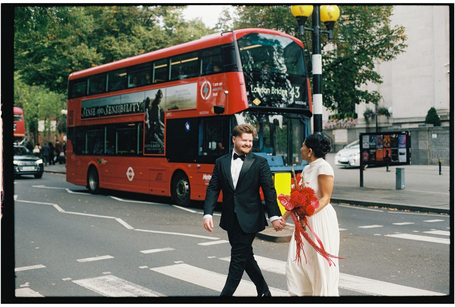 Couple in wedding attire crossing a zebra crossing in London with a red double-decker bus in the background.